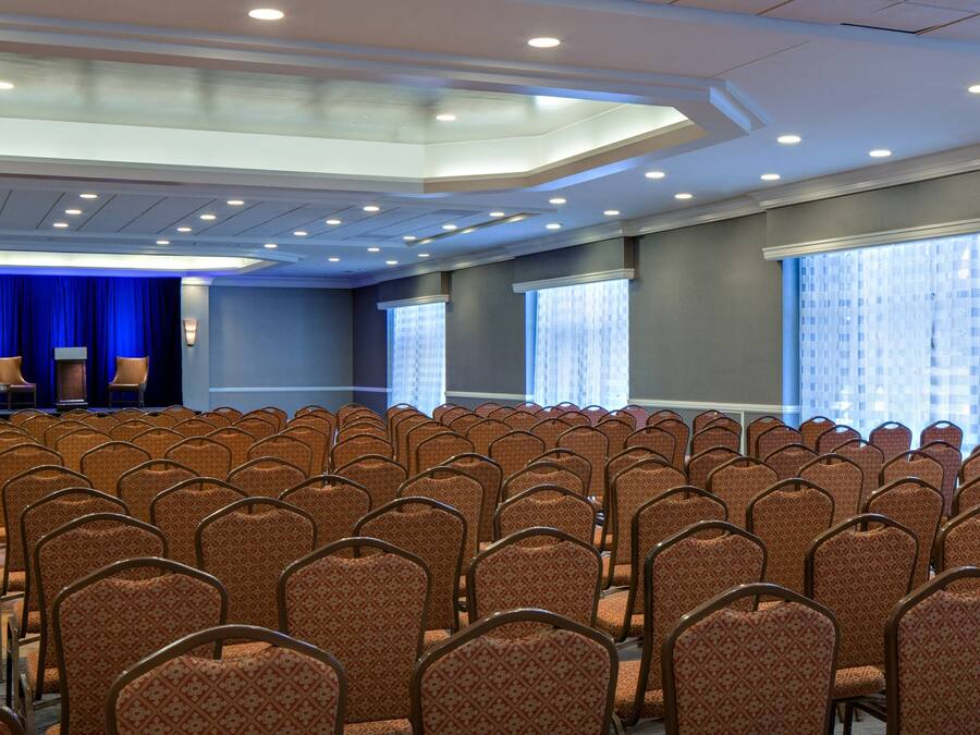 Un salón de conferencias con filas de sillas, un escenario con dos sillas y ventanas amplias con persianas.
