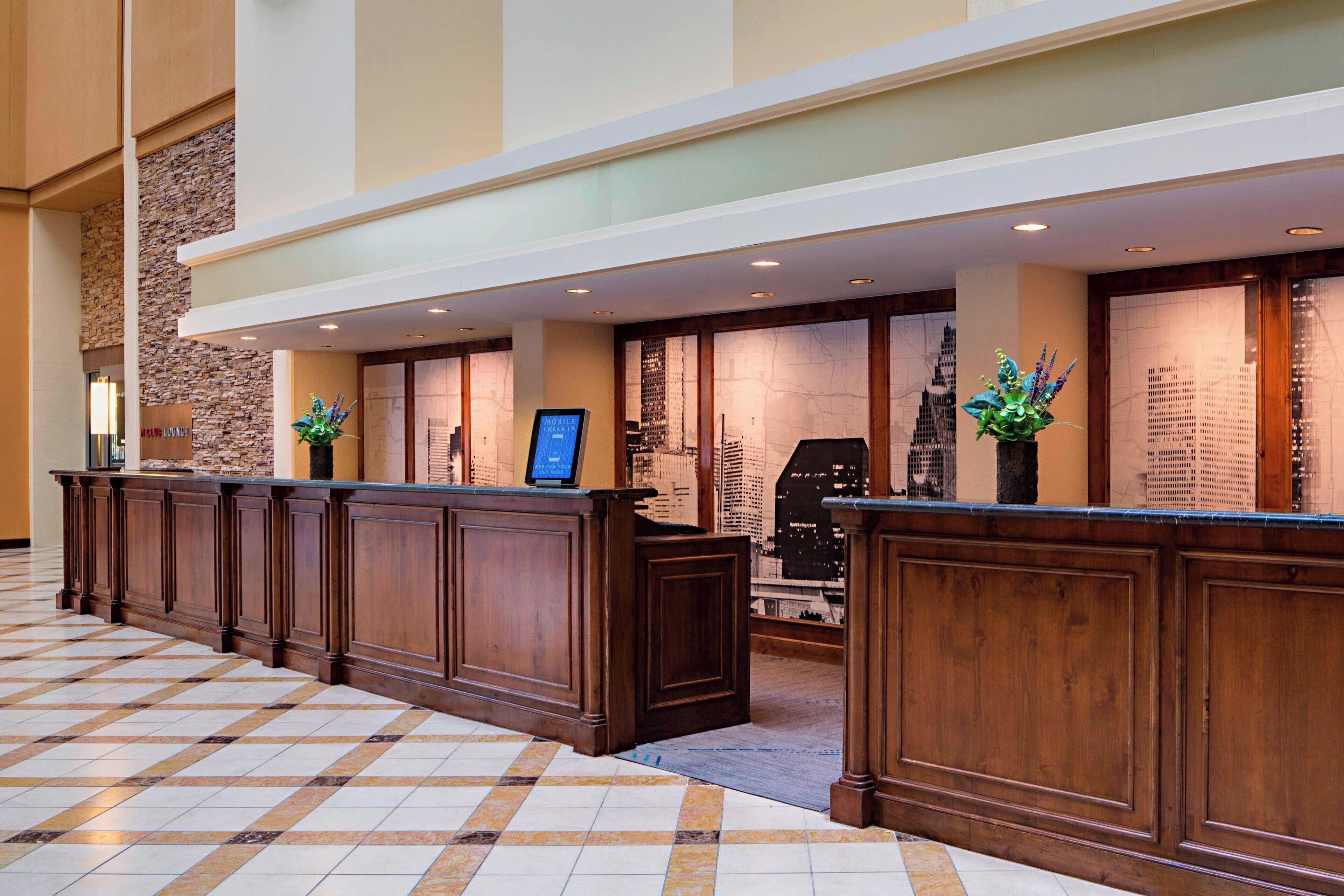 A hotel reception area with a long wooden reception desk, a stone accent wall, and a cityscape view through the windows.