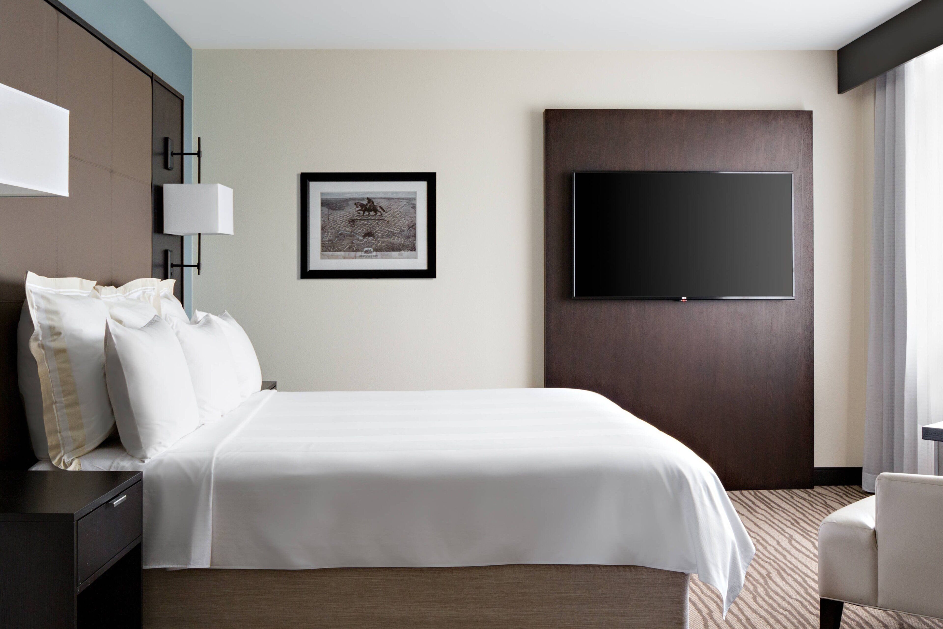 Quarto de hotel com uma cama grande, televisão de tela plana fixada na parede, um quadro emoldurado e uma escrivaninha com abajur.