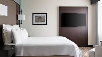 A hotel room with a large bed, a flat-screen TV mounted on the wall, a framed picture, and a desk with a lamp.