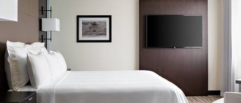 Quarto de hotel com uma cama grande, televisão de tela plana fixada na parede, um quadro emoldurado e uma escrivaninha com abajur.