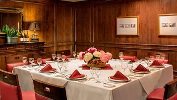 Une salle à manger avec une longue table dressée pour un repas, des chaises à revêtement rouge et un arrangement floral en guise de centre de table.
