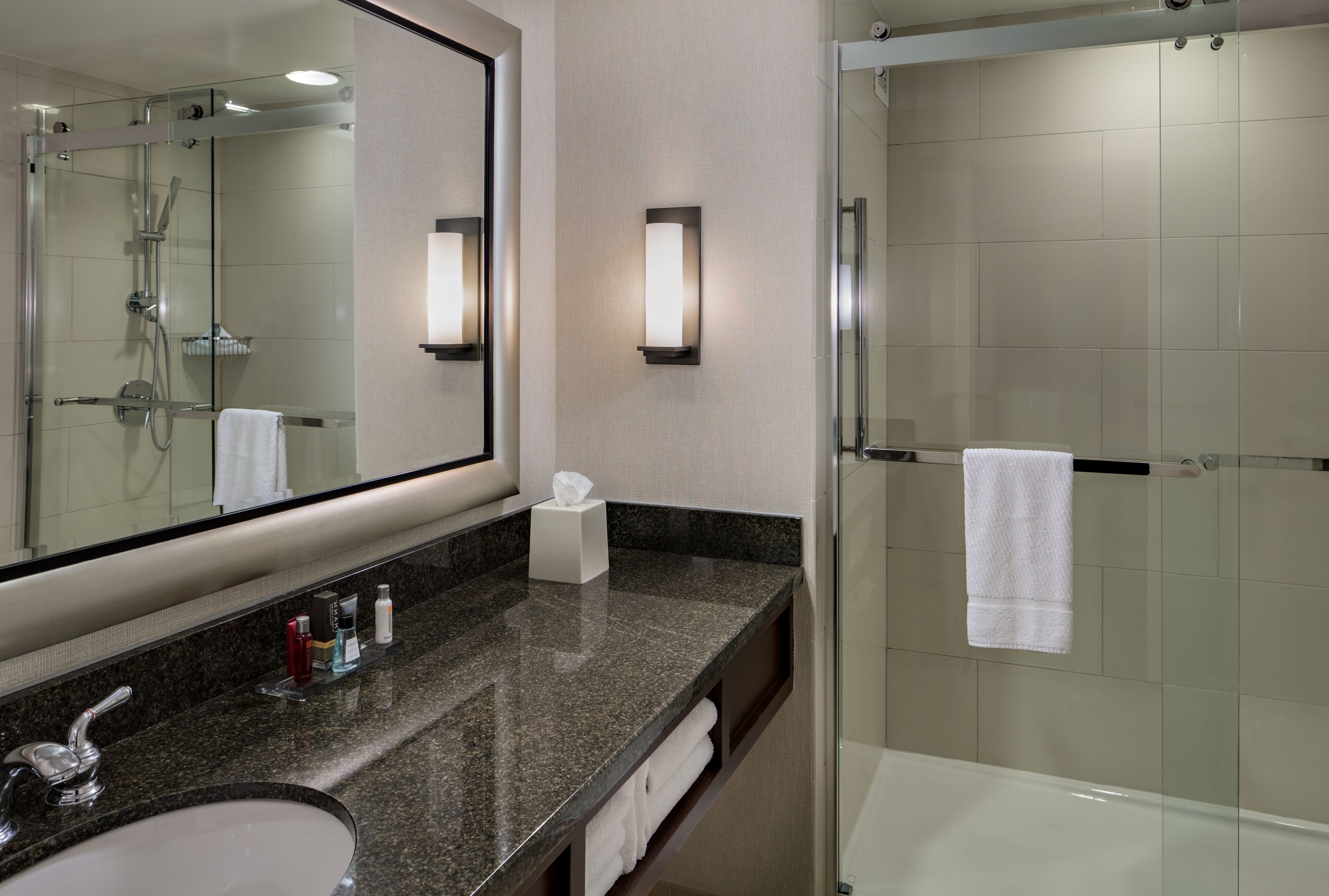 A modern bathroom with a granite countertop, a large mirror, a glass-enclosed shower, and a white bathtub.