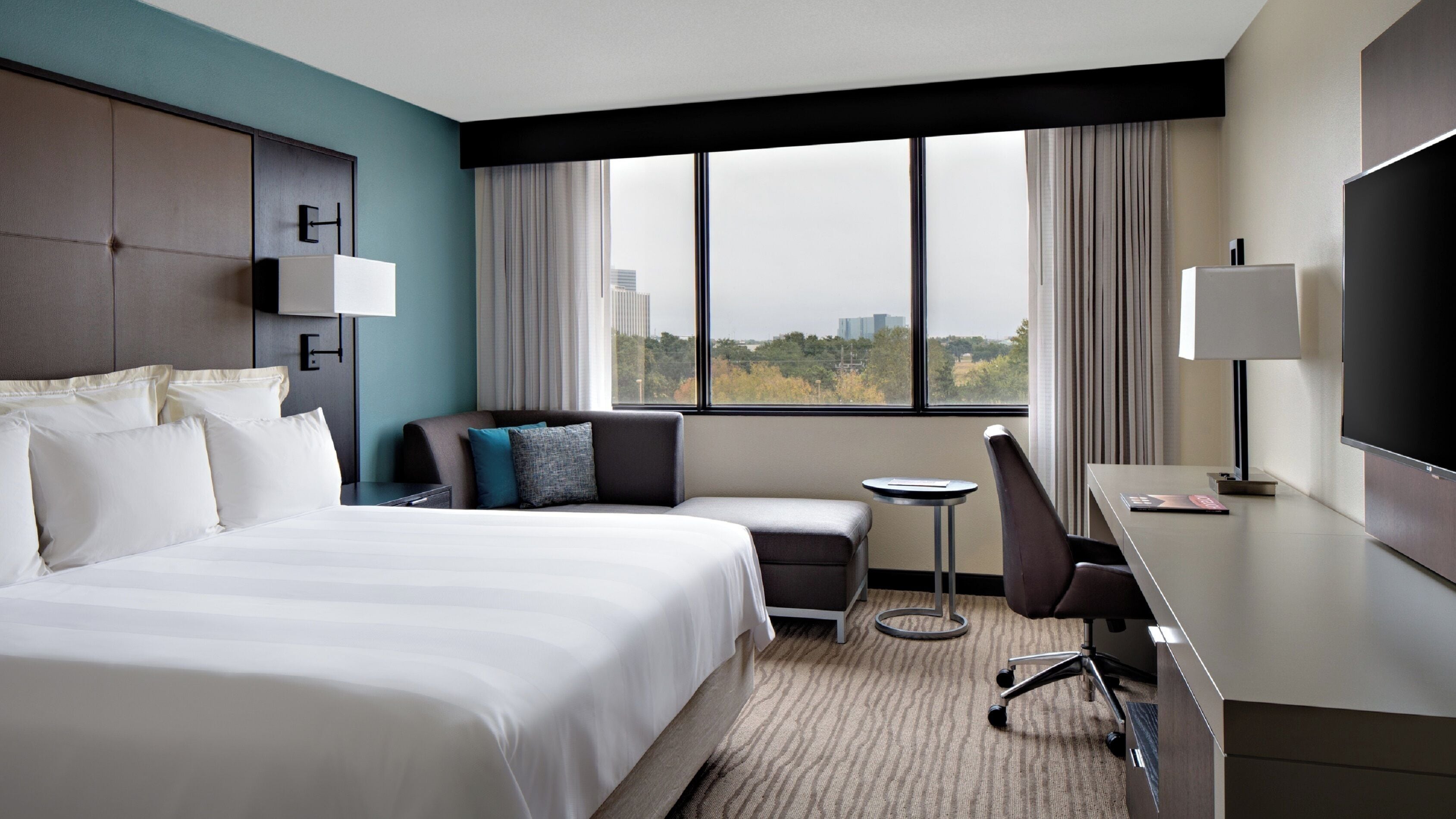 A hotel room with a large bed, a desk, a chair, and a view of the city through the window.