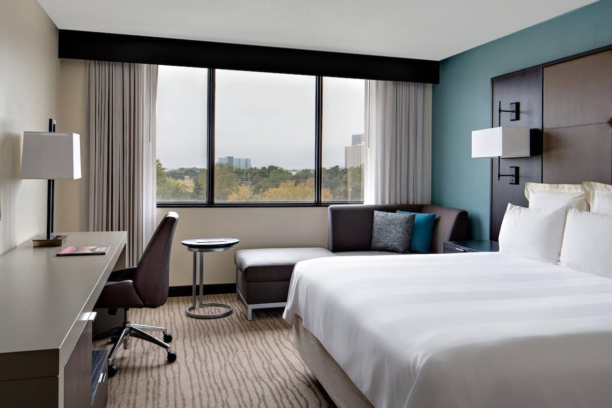 A hotel room with a bed, a desk, a chair, a small table, and a view of the city through large windows.