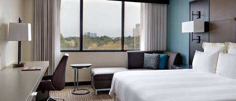 A hotel room with a bed, a desk, a chair, a small table, and a view of the city through large windows.