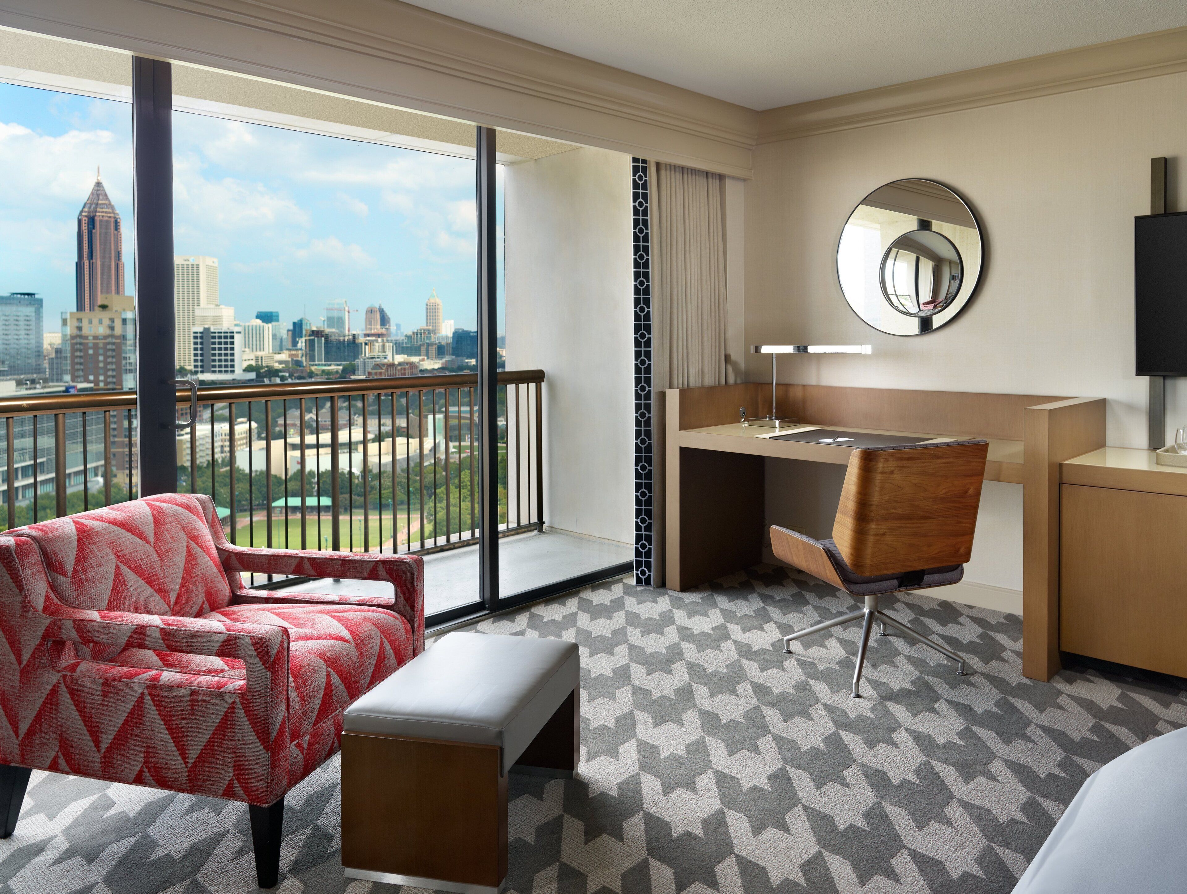 A hotel room with a balcony, a desk, a chair, a bench, and a large window offering a city view.