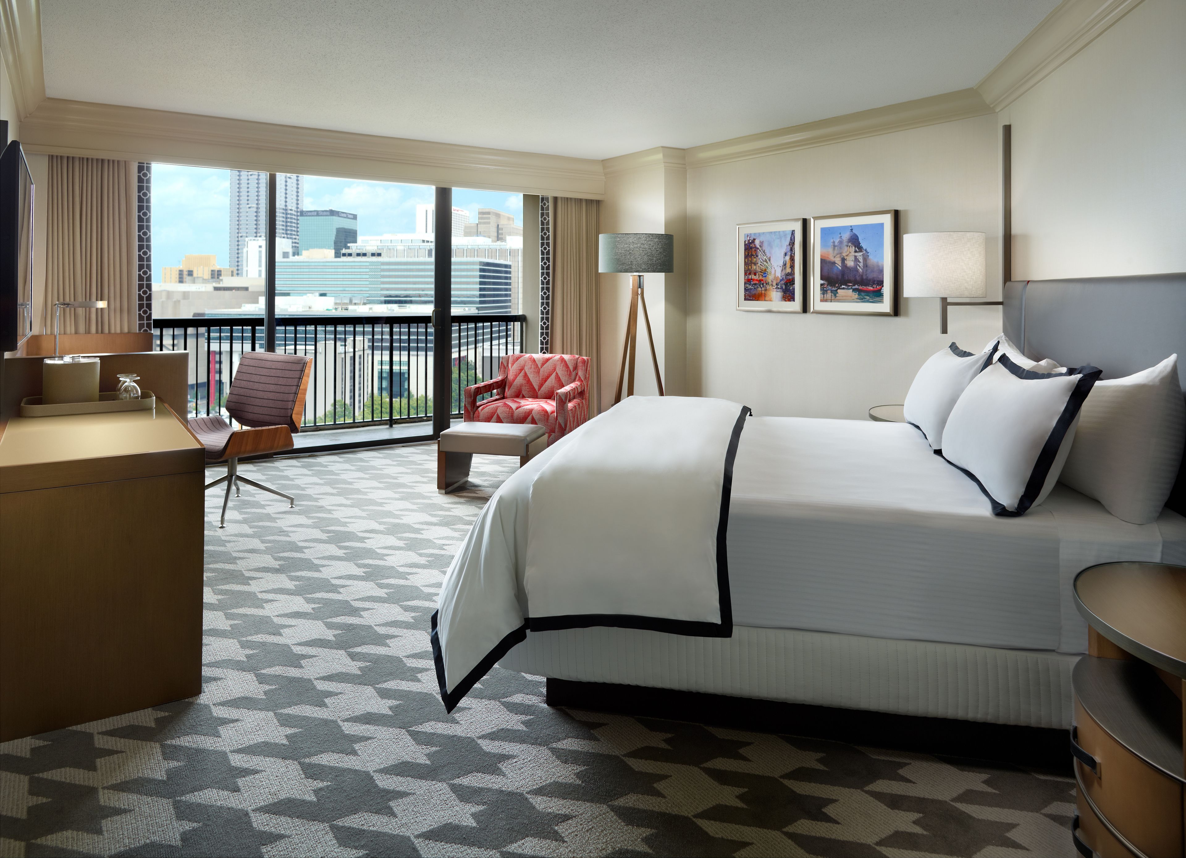 A hotel room with a large bed, a desk, a chair, a balcony with a view of the city, and two framed pictures on the wall.