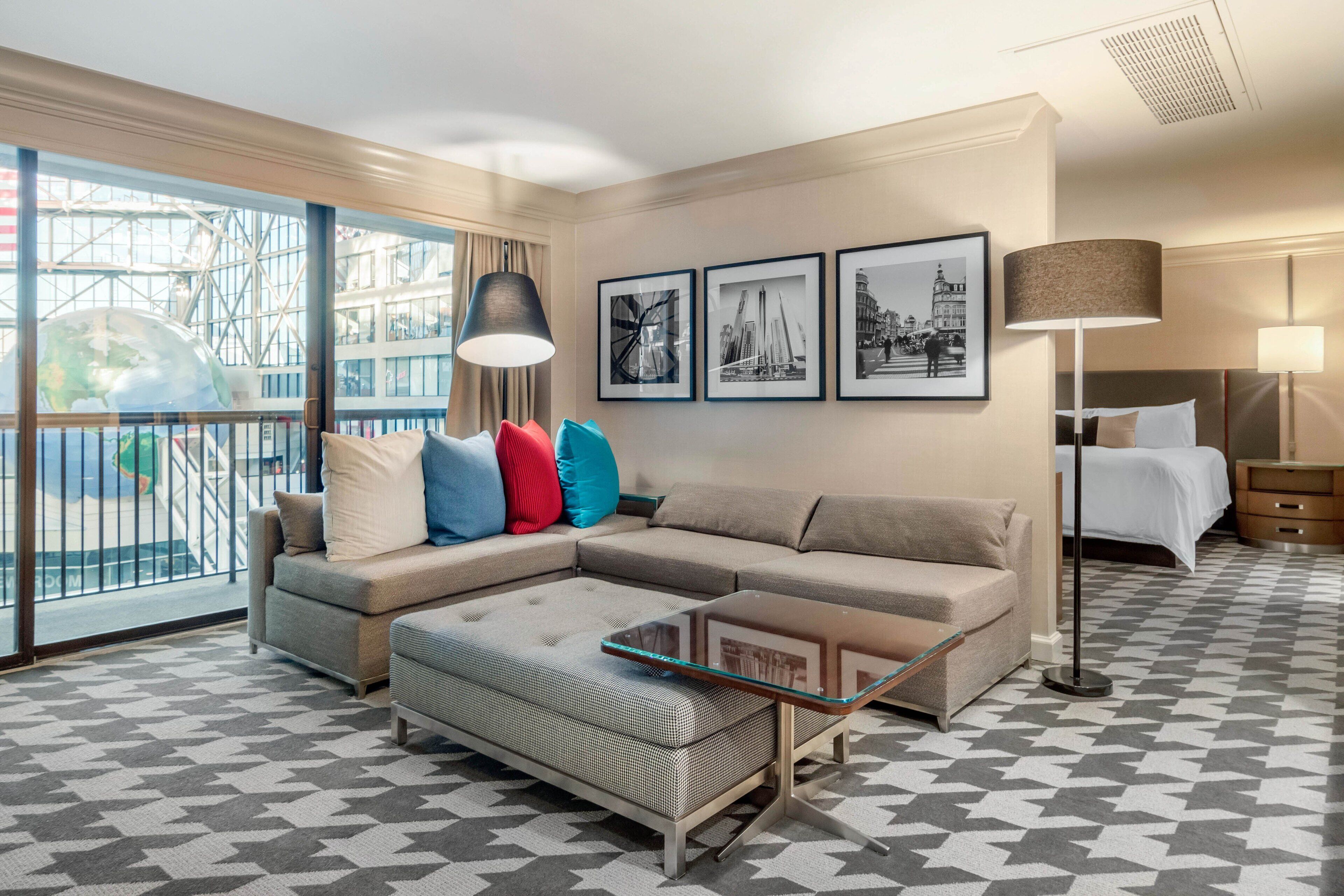 A modern hotel room with a large window, a sofa, a glass coffee table, and a bed.