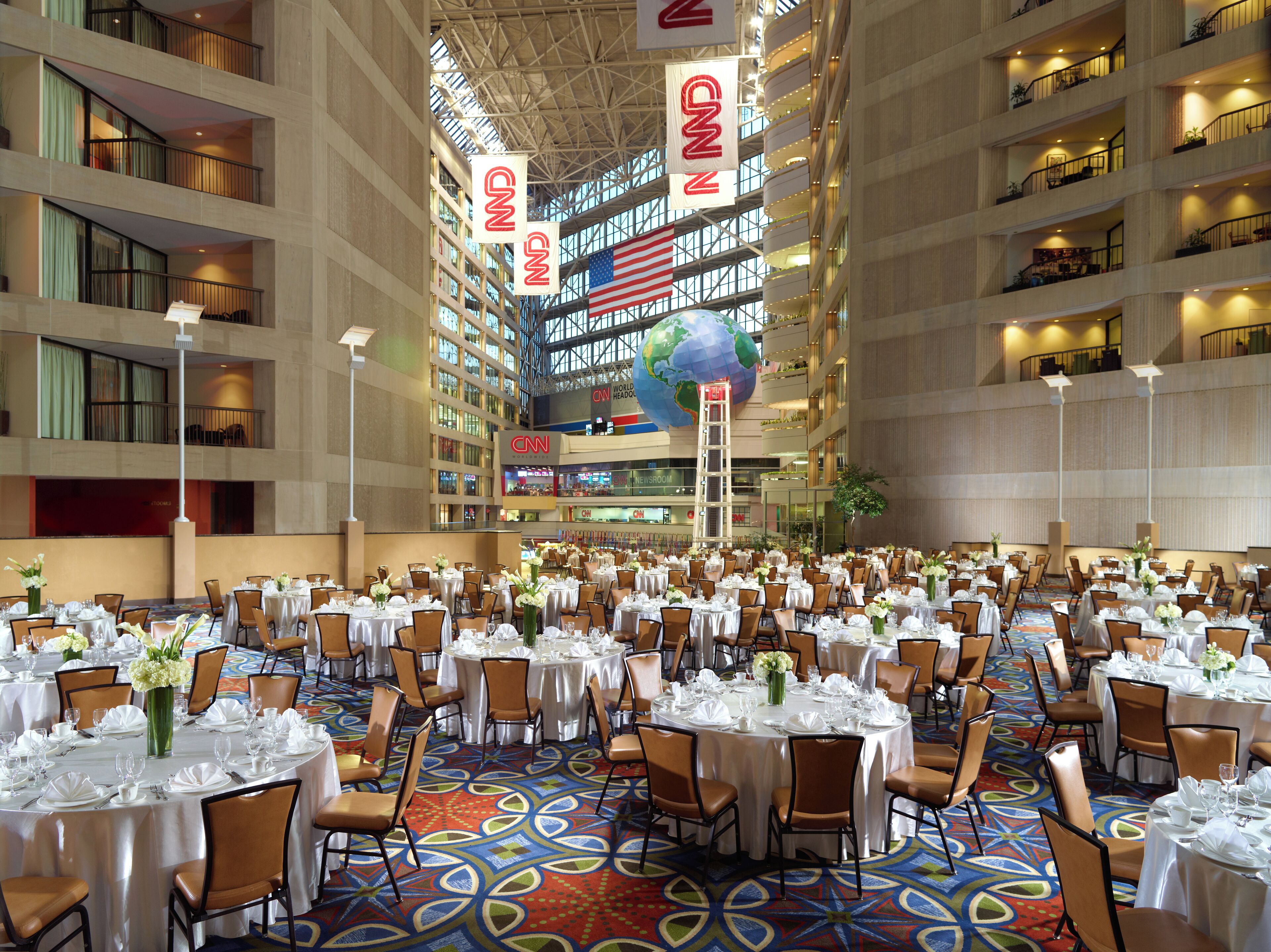 A large indoor banquet hall with multiple round tables set for dining, a CNN World Headquarters sign, and a large American flag.