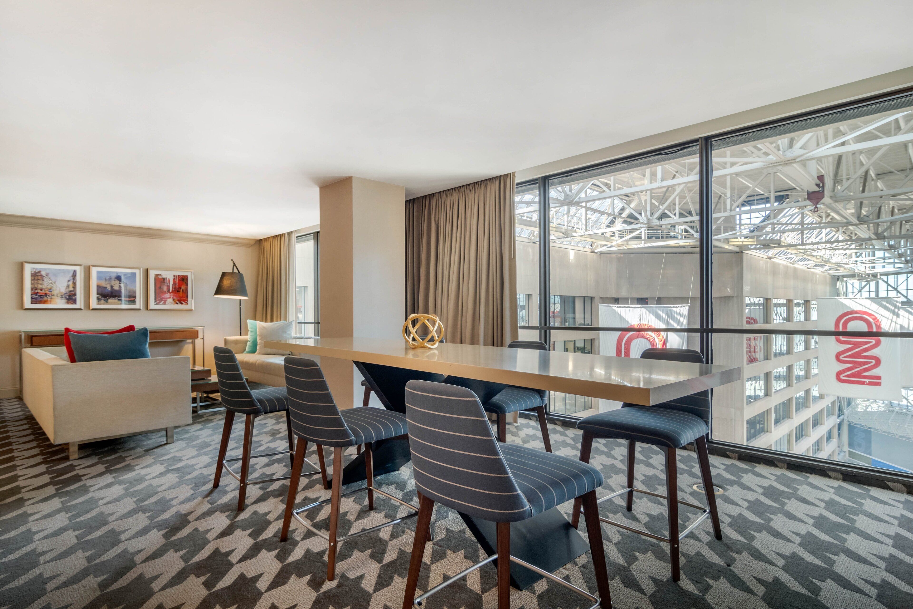Una habitación de hotel moderna con un ventanal grande, una zona de comedor con una mesa larga y sillas, un sofá y una alfombra estampada.