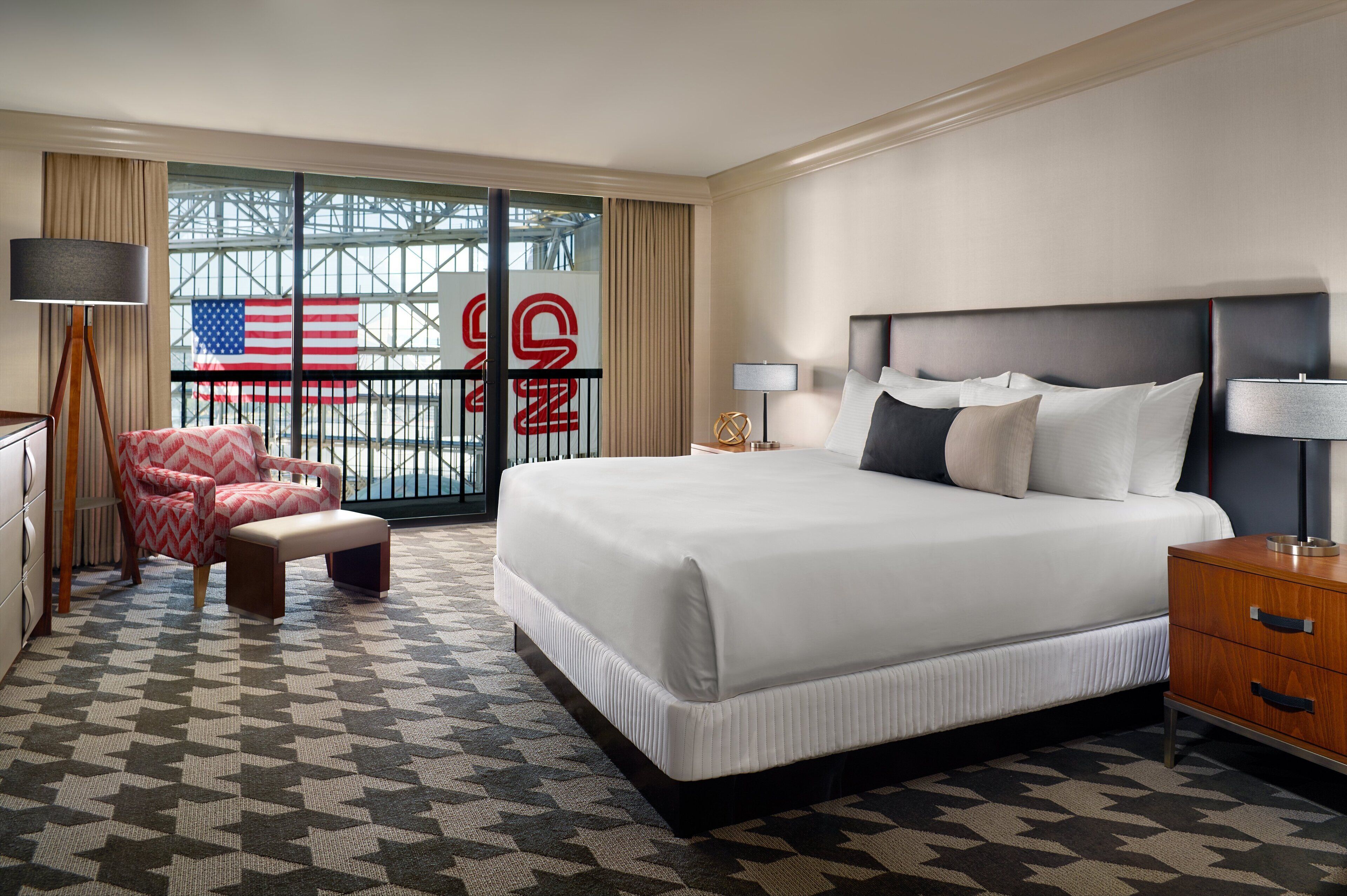 A hotel room with a large bed, a chair, a nightstand, and a view of the American flag through a balcony door.