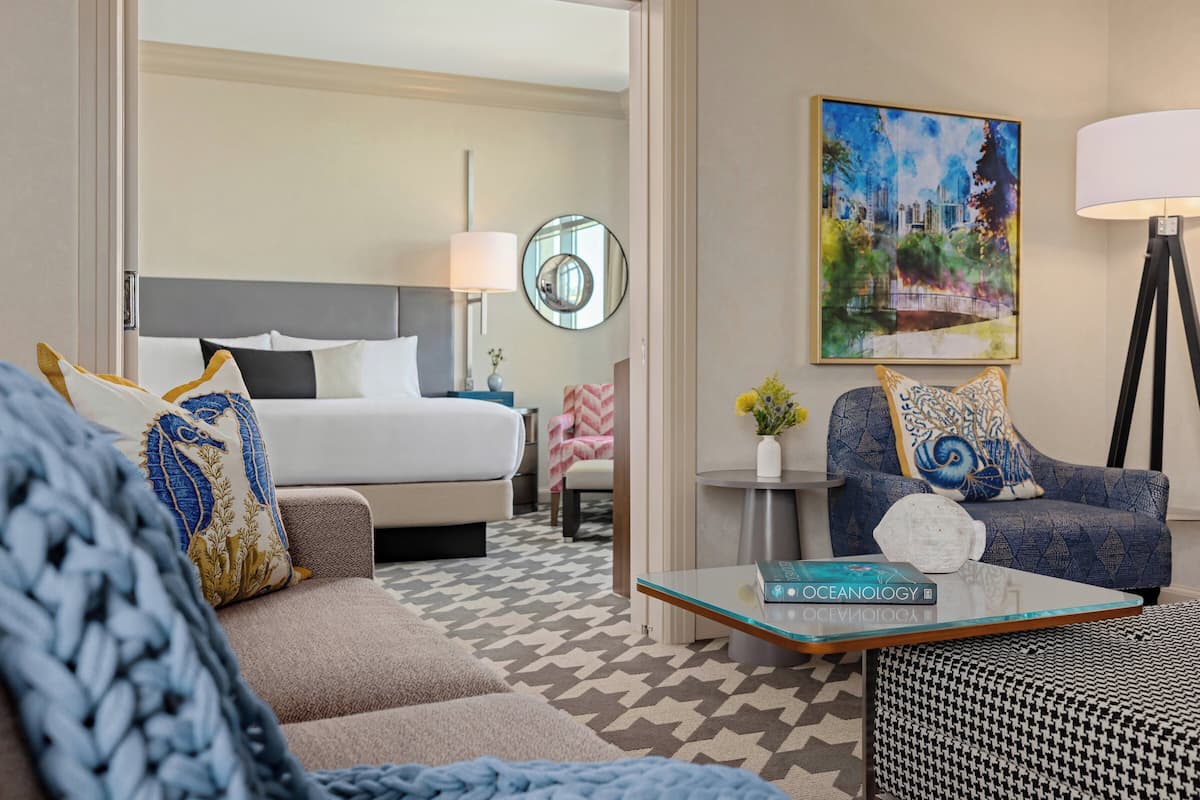 A hotel room with a bed, a sofa, a coffee table, a chair, a lamp, and a painting.