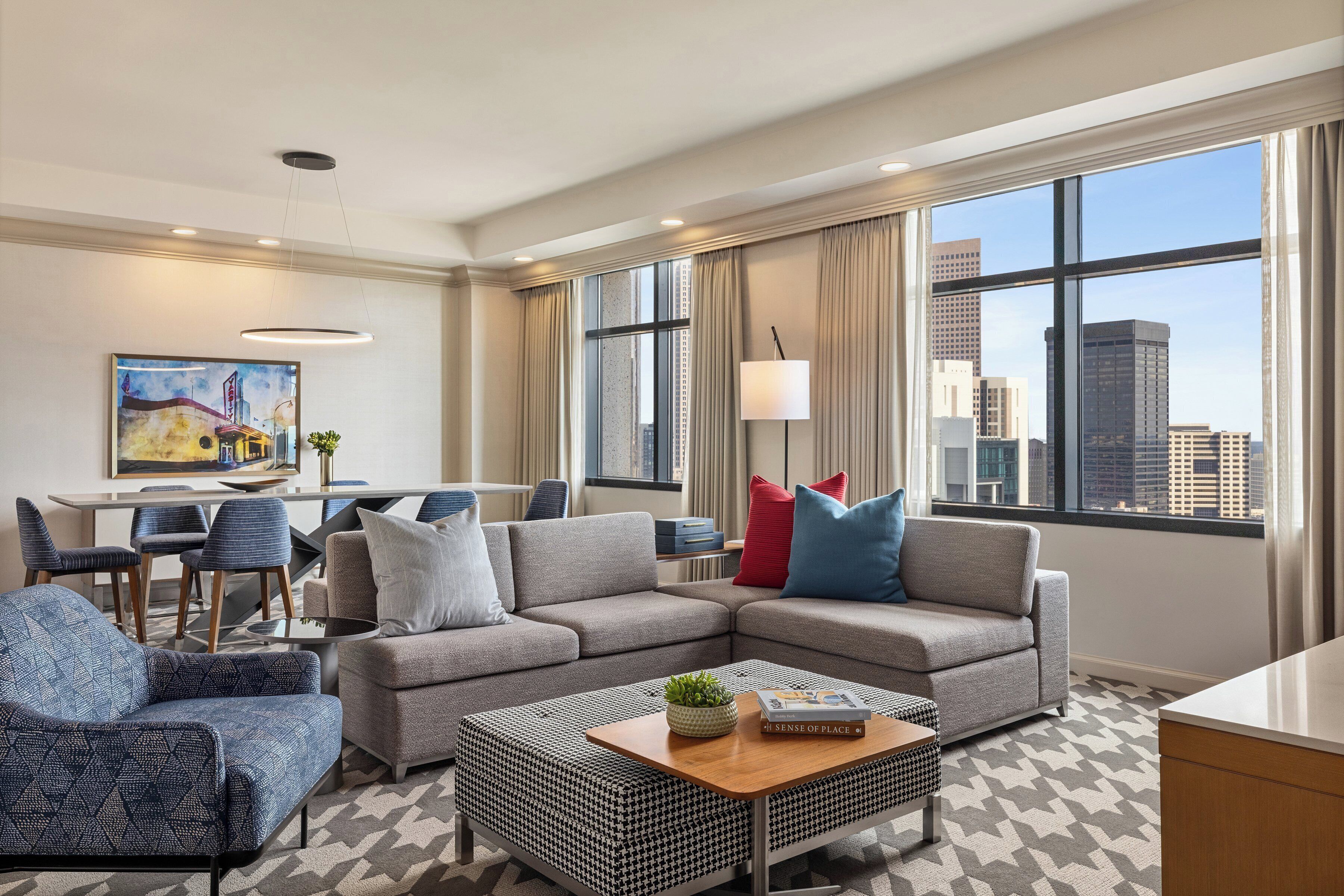 A modern hotel room with a sectional sofa, a dining area, and a large window with a city view.