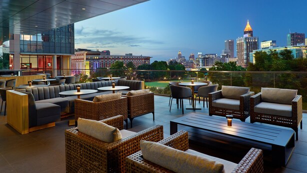 An outdoor seating area with wicker furniture and tables, overlooking a cityscape with buildings and a stadium. - Omni Atlanta Hotel at Centennial Park (Atlanta)