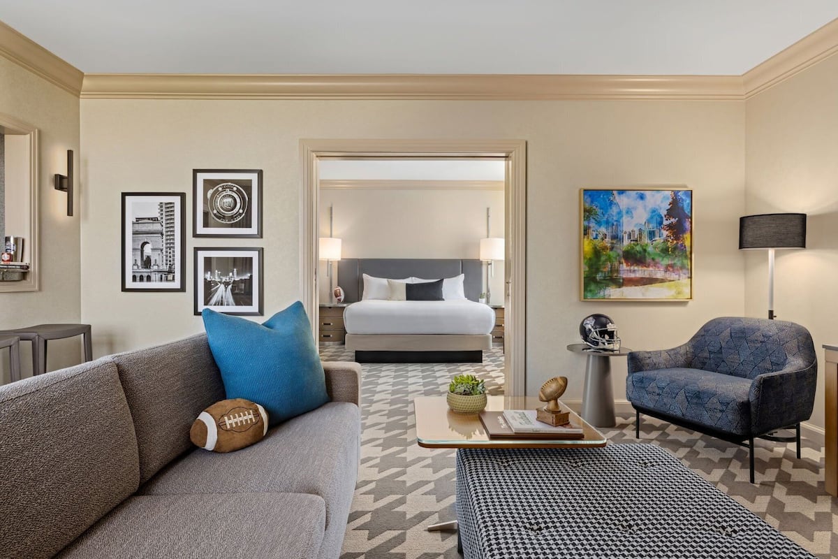 A hotel room with a large bed, a sofa, a coffee table, and a chair. There are framed pictures on the wall and a painting above the bed.