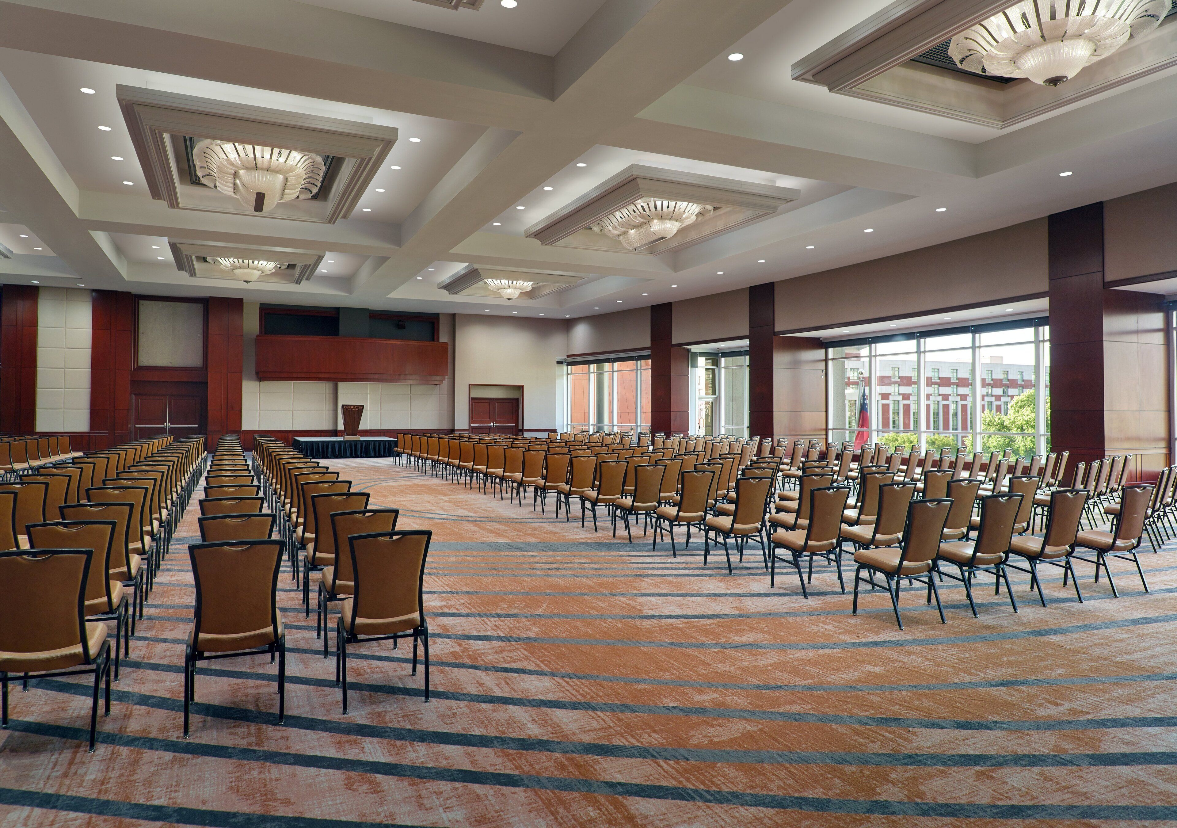 A large conference room with rows of chairs and a stage, featuring a high ceiling with recessed lighting and large windows.