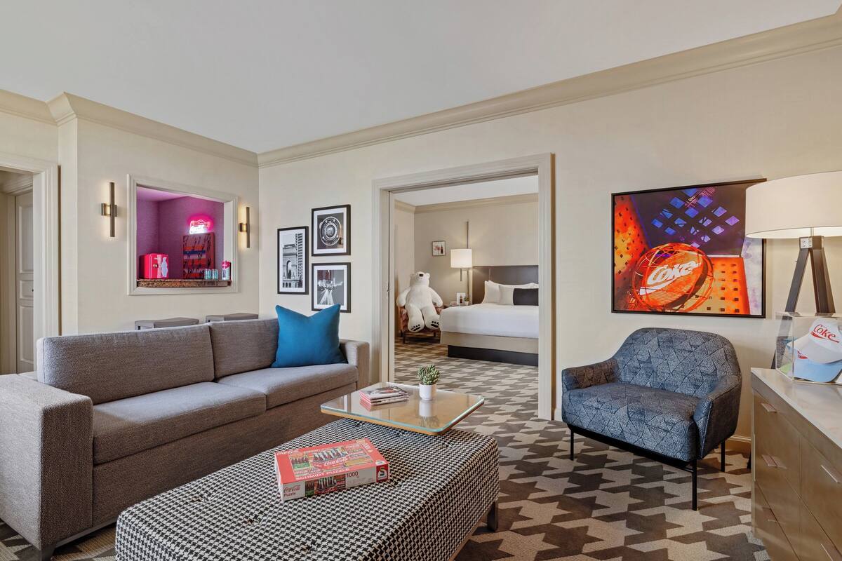 A modern hotel room with a sofa, armchair, and a coffee table with a book. There is a bed with a headboard and a large painting on the wall.