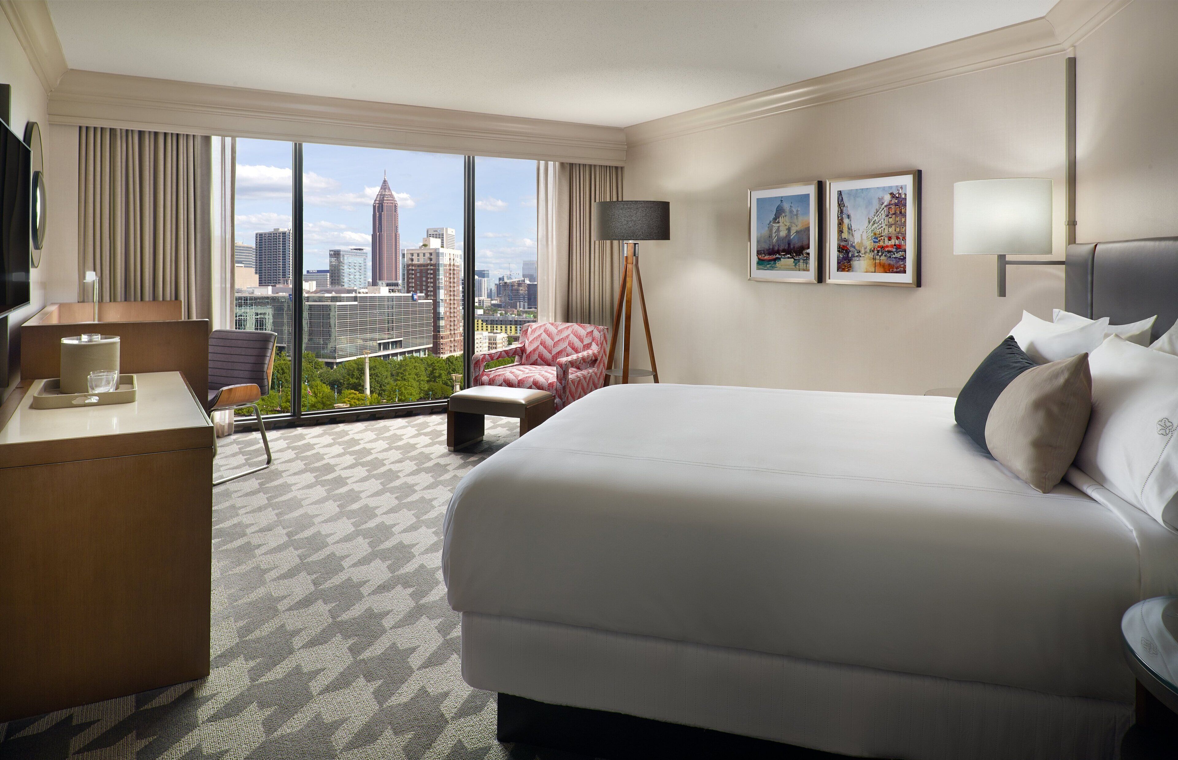 A hotel room with a large bed, a desk, a chair, a floor lamp, and a city view.