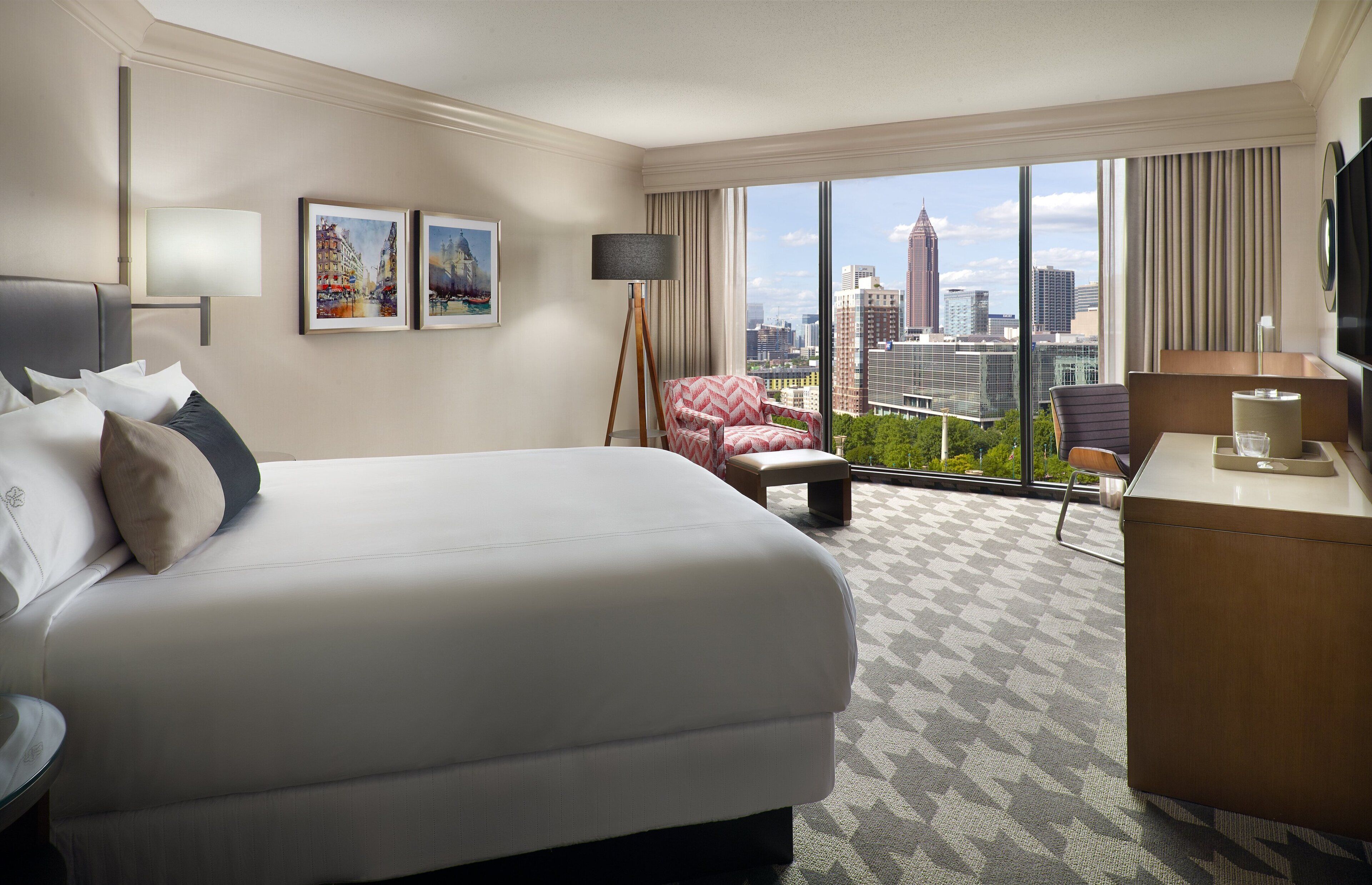 A hotel room with a large bed, a desk, a chair, a lamp, and a view of the cityscape through large windows.