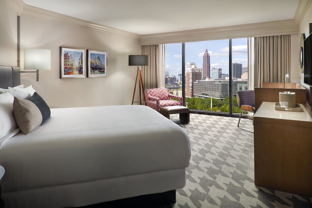 A hotel room with a large bed, a desk, a chair, a lamp, and a view of the cityscape through large windows.