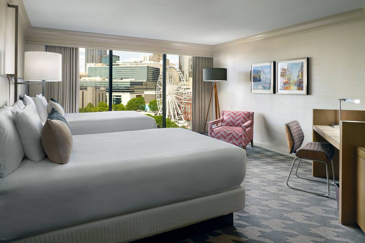 A hotel room with two beds, a desk, a chair, a lamp, and a large window with a city view.