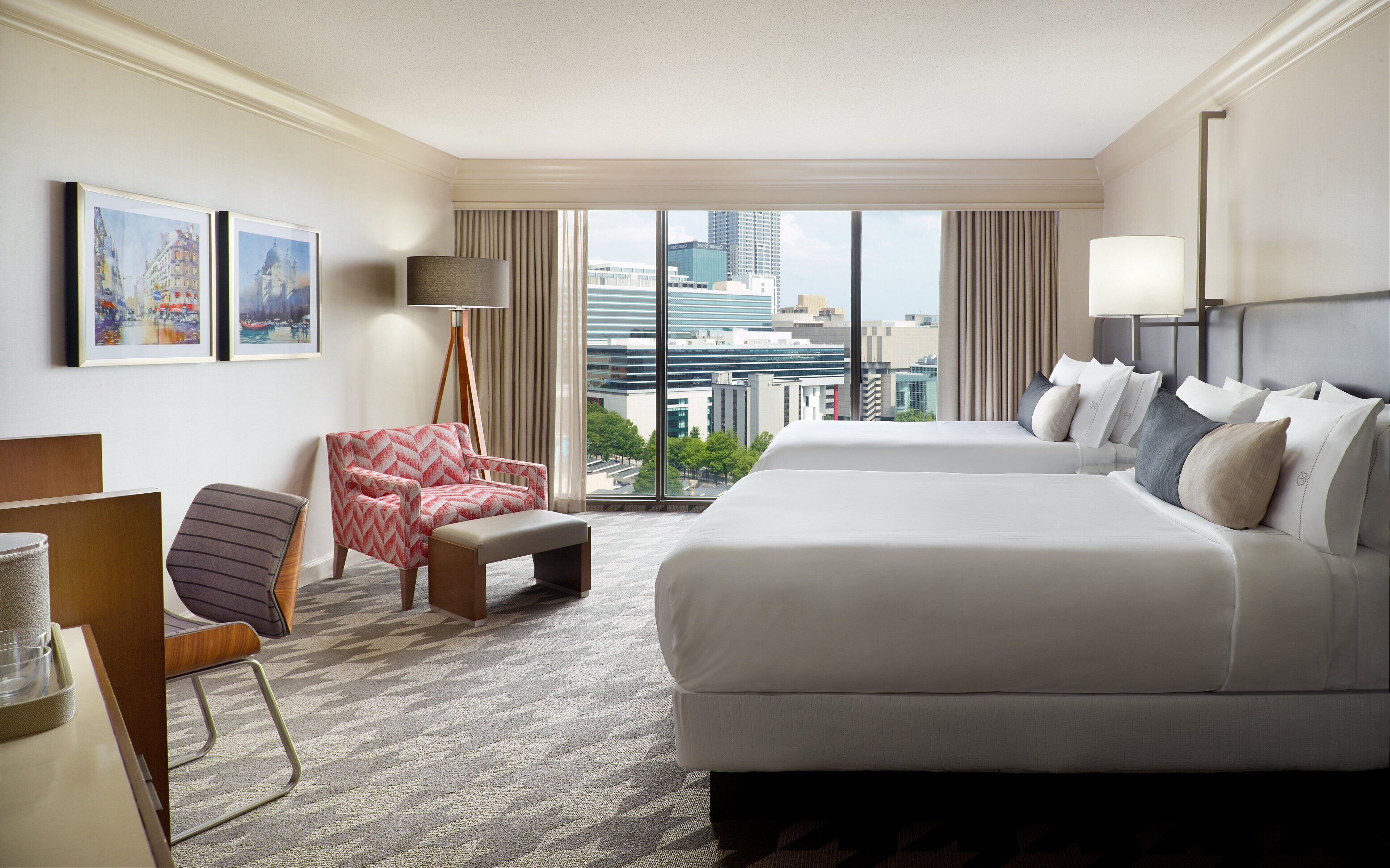 A hotel room with a large bed, a chair, a desk, and a view of the cityscape through the window.