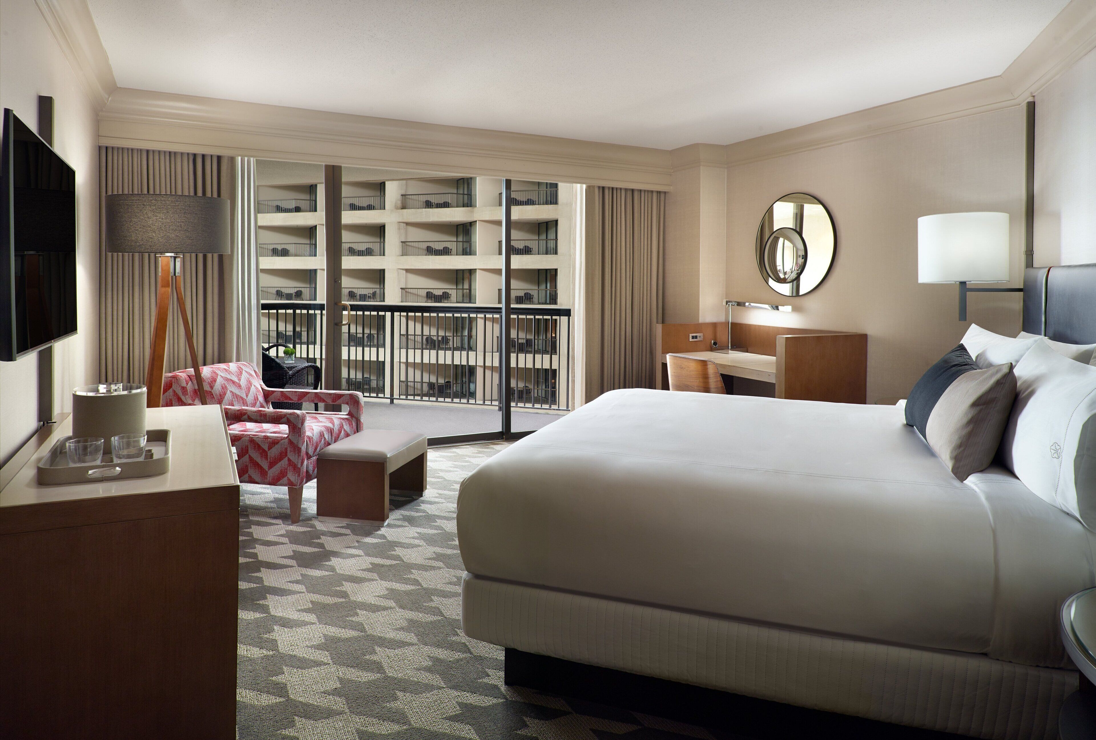 A hotel room with a large bed, a desk with a lamp, a TV, and a balcony with a view of the building's interior.