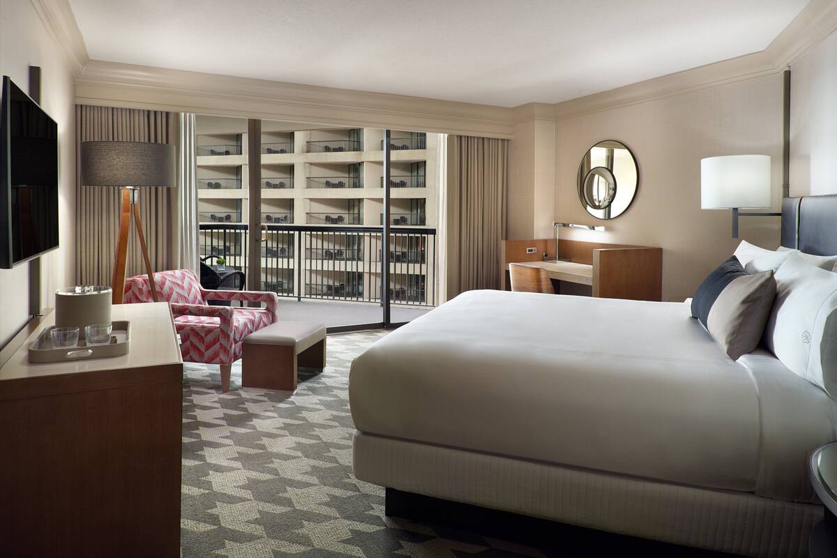 A hotel room with a large bed, a desk with a lamp, a TV, and a balcony with a view of the building's interior.