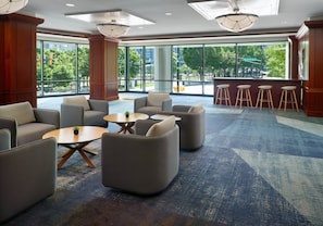 A spacious lobby with modern furniture, large windows offering a view of the outdoors, and a bar area with stools.