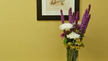 Una obra de arte enmarco sobre una pared amarilla, un jarrón con flores sobre una mesa de madera.