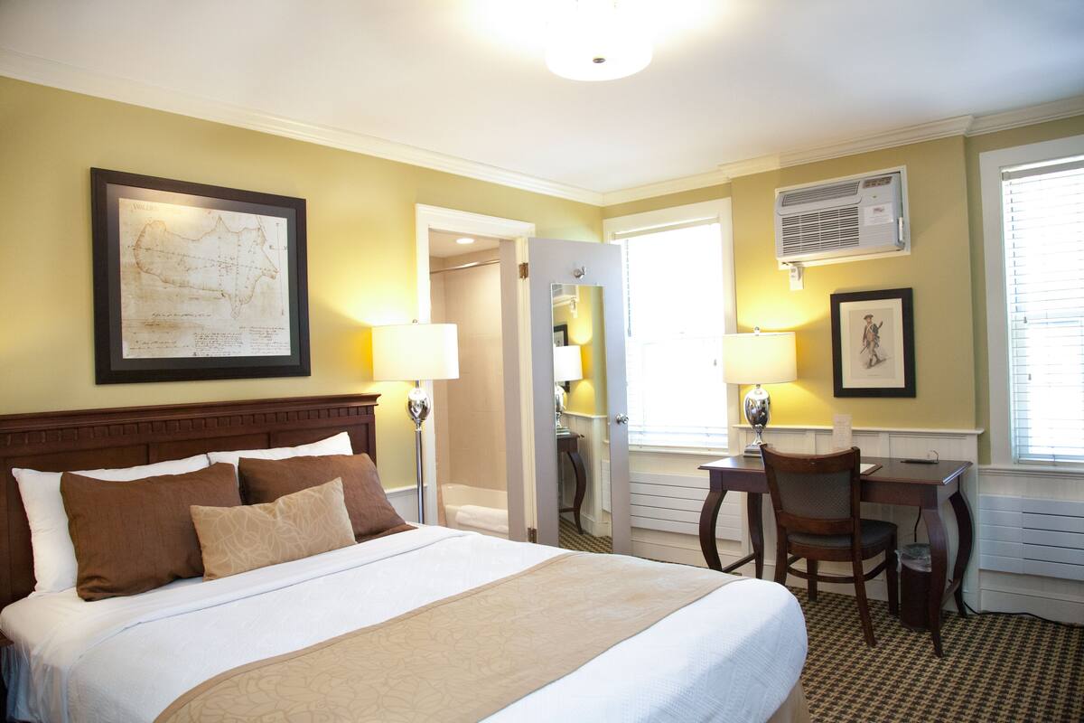 Quarto de hotel com uma cama grande, duas mesas de cabeceira com abajures, uma escrivaninha com cadeira, um mapa emoldurado na parede e um aparelho de ar condicionado.