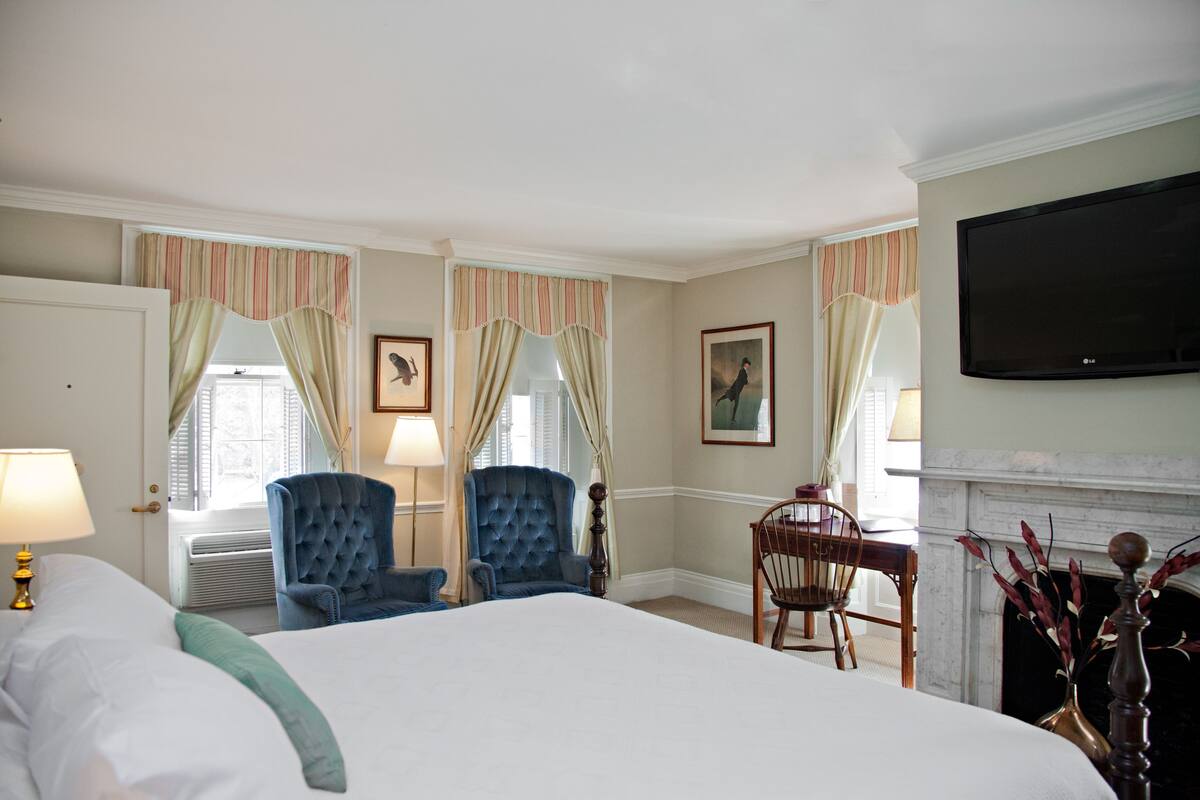 Quarto de hotel com uma cama grande, duas poltronas azuis, uma cadeira de madeira, lareira e televisão de tela plana.