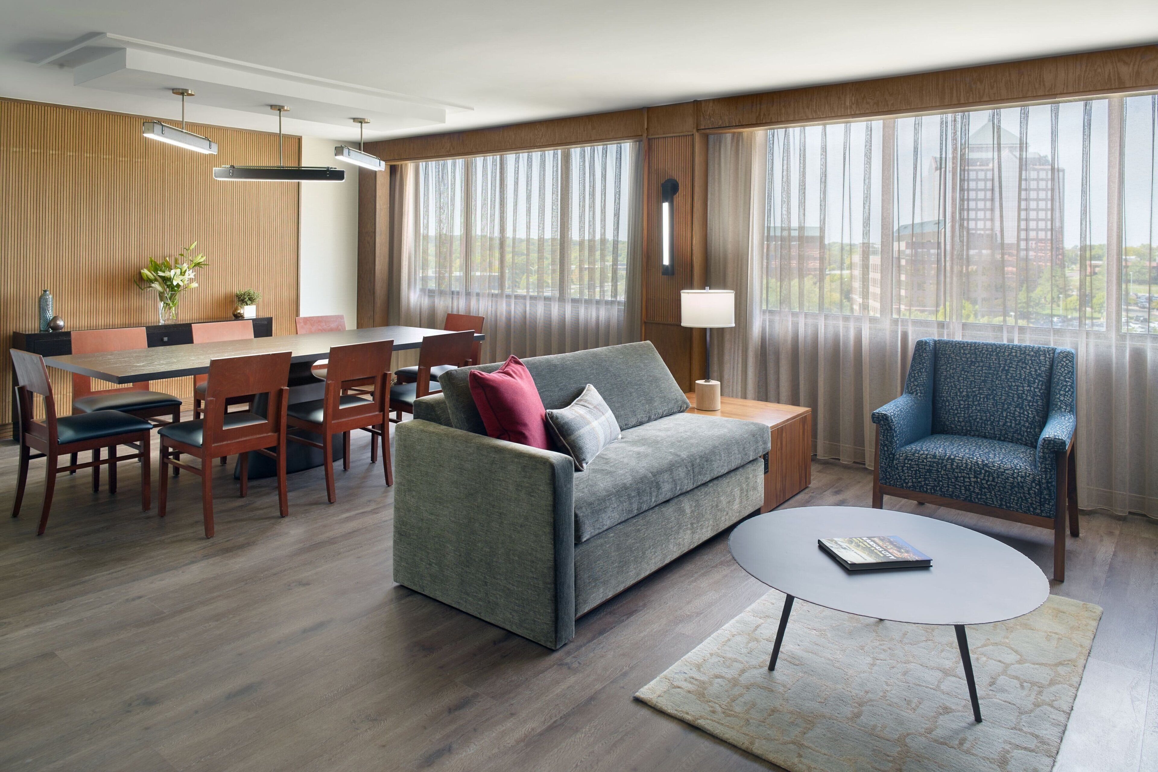 A modern hotel room with a dining area, a sofa, a coffee table, and large windows with a city view.