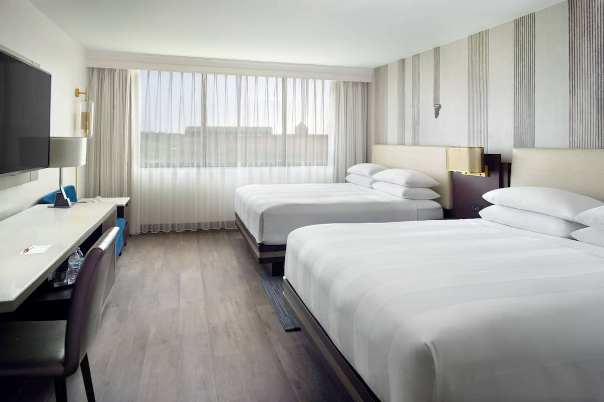 Ein Hotelzimmer mit zwei Betten, einem Schreibtisch, einem Sessel, einem Fernseher und einem großen Fenster mit Vorhängen.