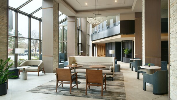Un hall d’entrée spacieux avec un plafond haut, de grandes fenêtres, un espace salon central avec un canapé et des fauteuils, et un escalier menant à un étage supérieur.
