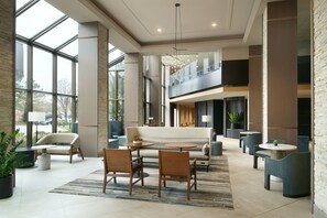 Un hall d’entrée spacieux avec un plafond haut, de grandes fenêtres, un espace salon central avec un canapé et des fauteuils, et un escalier menant à un étage supérieur.