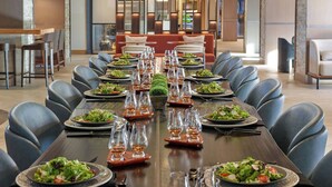 Une longue table à manger, dressée avec des assiettes de salade, des verres à vin et des couverts, est entourée de chaises bleues dans un espace de repas moderne. On aperçoit en arrière-plan un canapé et des panneaux muraux décoratifs.