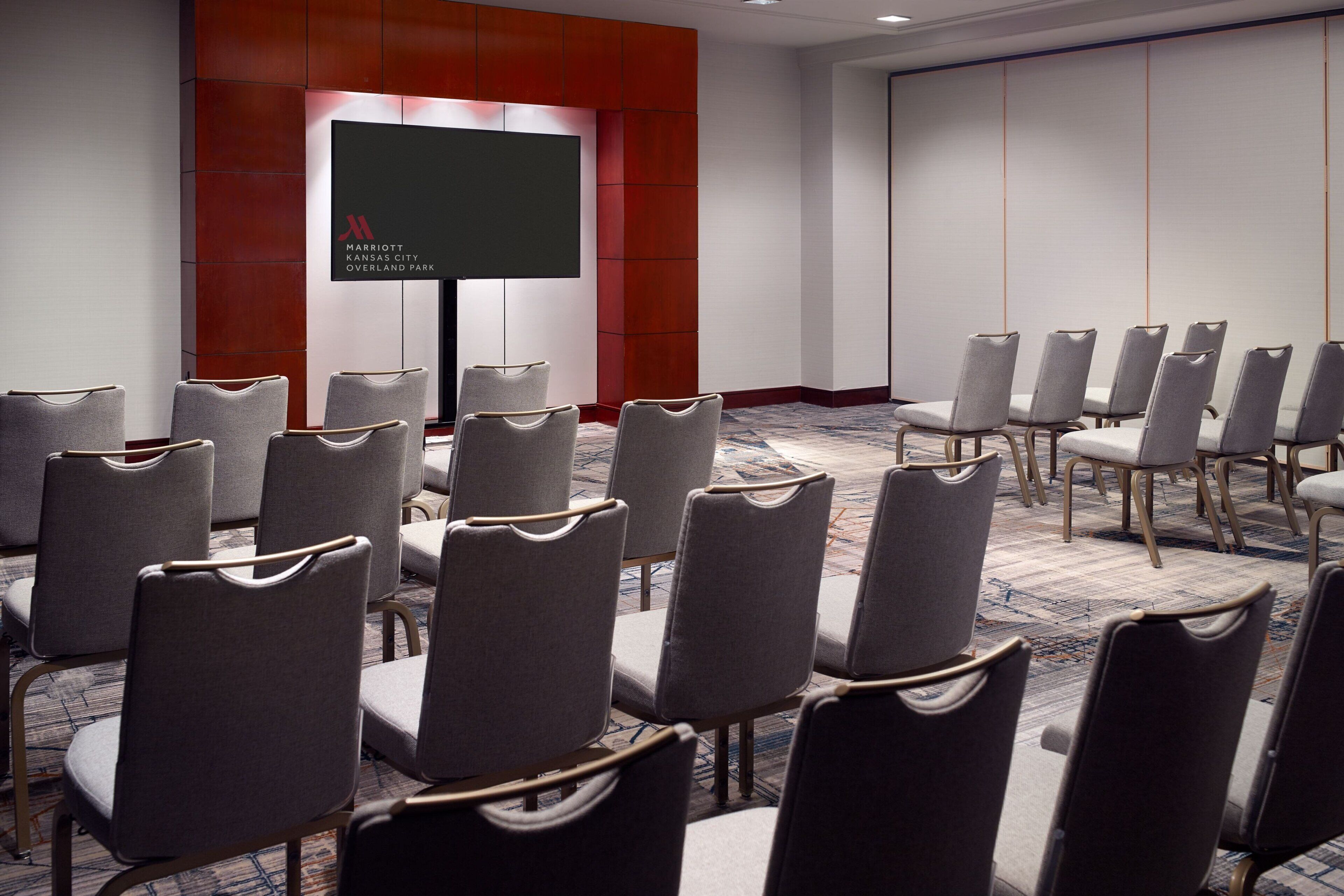 Un salón de conferencias con filas de sillas grises, una pantalla grande y una pared de acento roja.