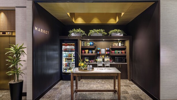 Un interior moderno con una máquina expendedora, estantes llenos de aperitivos, una mesa con vasos y una planta en maceta.