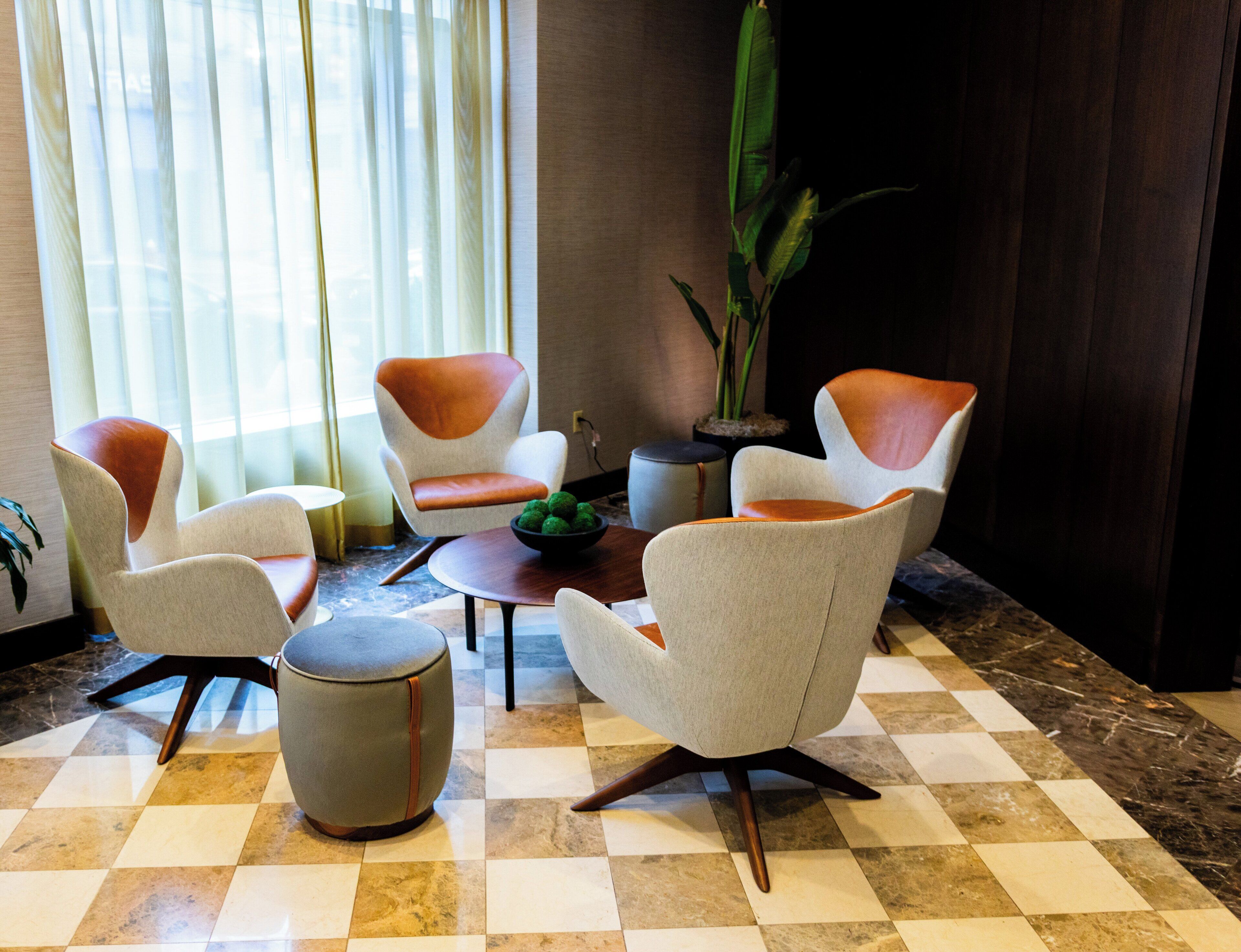 A modern lobby with checkered flooring, contemporary armchairs, a round table with a green centerpiece, and a tall potted plant.