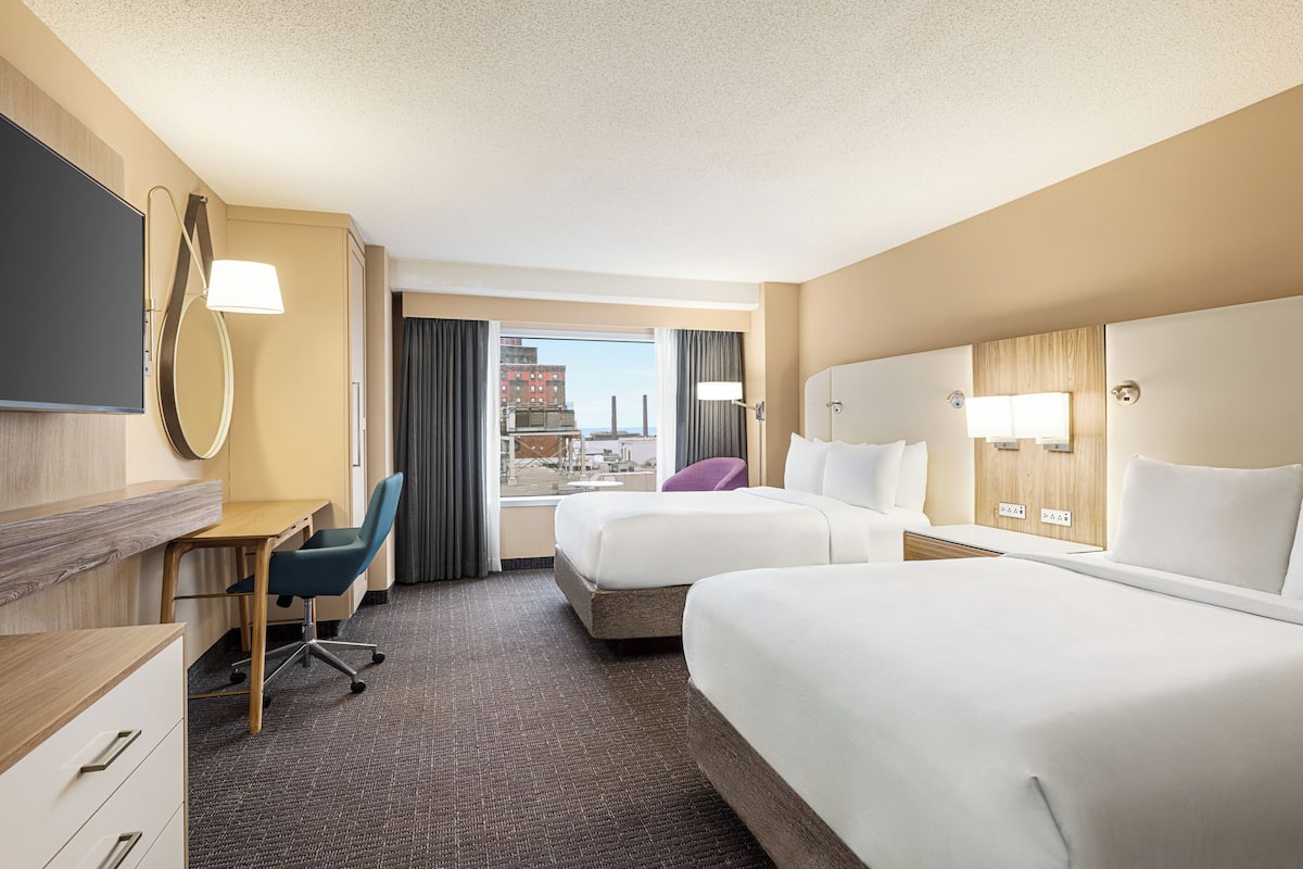A hotel room with two beds, a desk, a chair, a TV, and a view of the city.