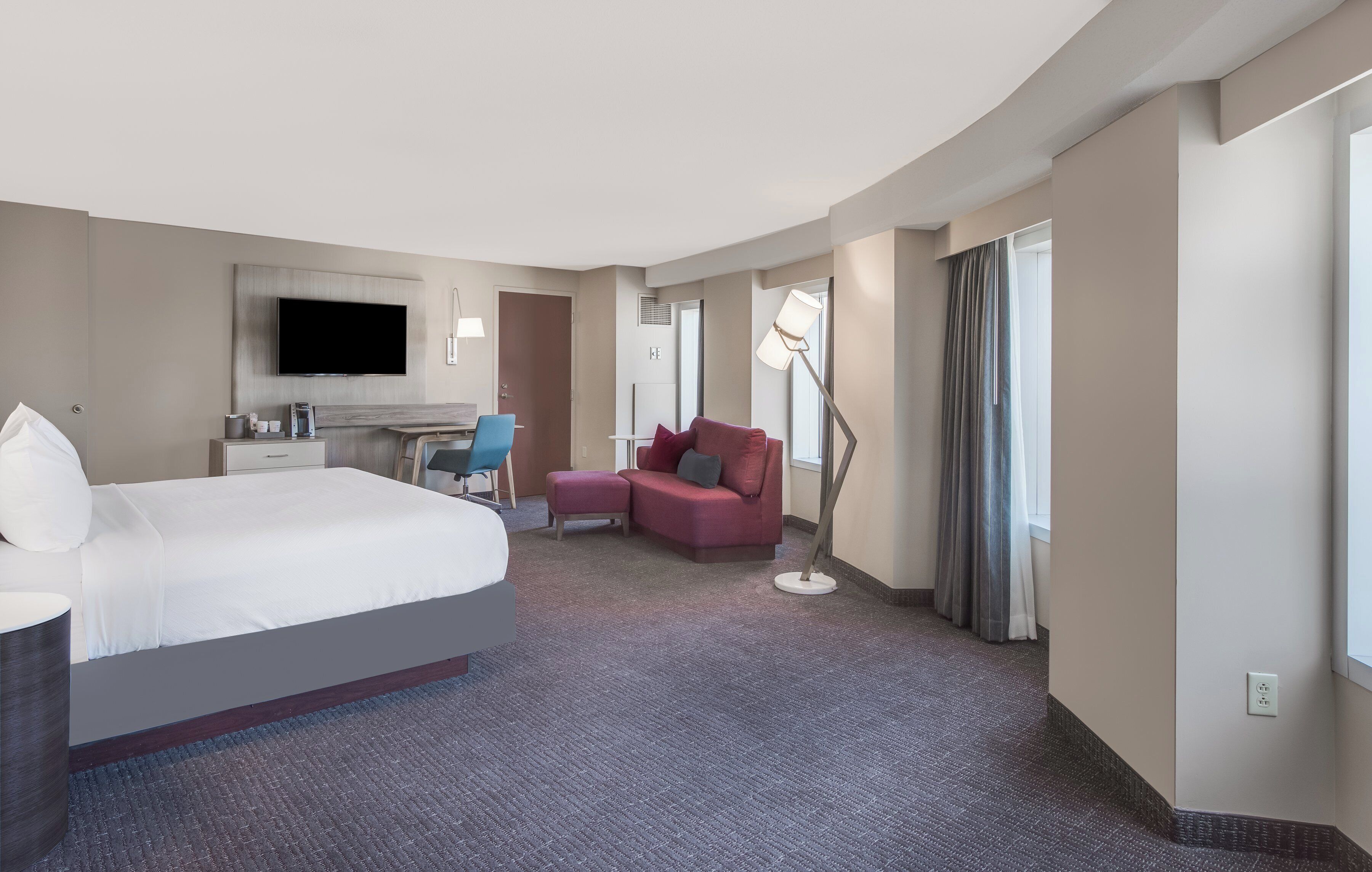 A hotel room with a large bed, a desk with a chair, a sofa, a TV, and a floor lamp.