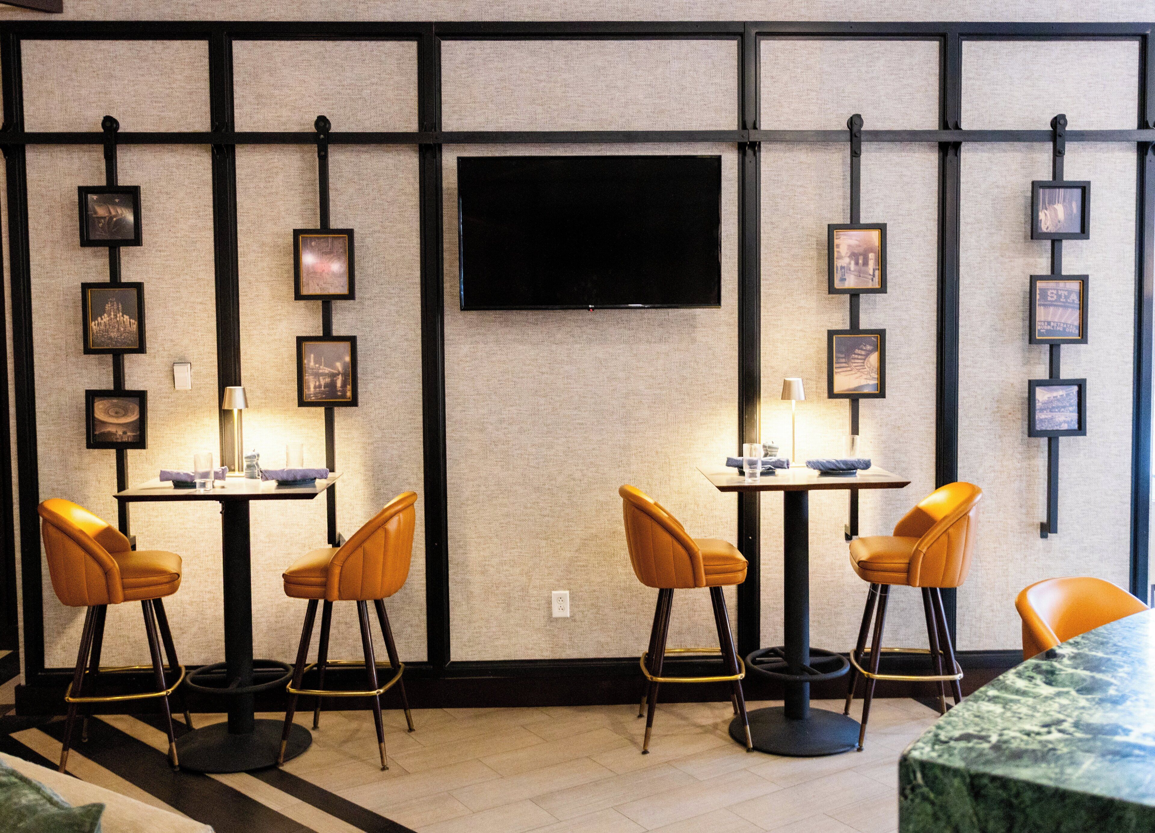 A hotel room with a TV mounted on the wall, two tables with chairs, and framed pictures on the wall.