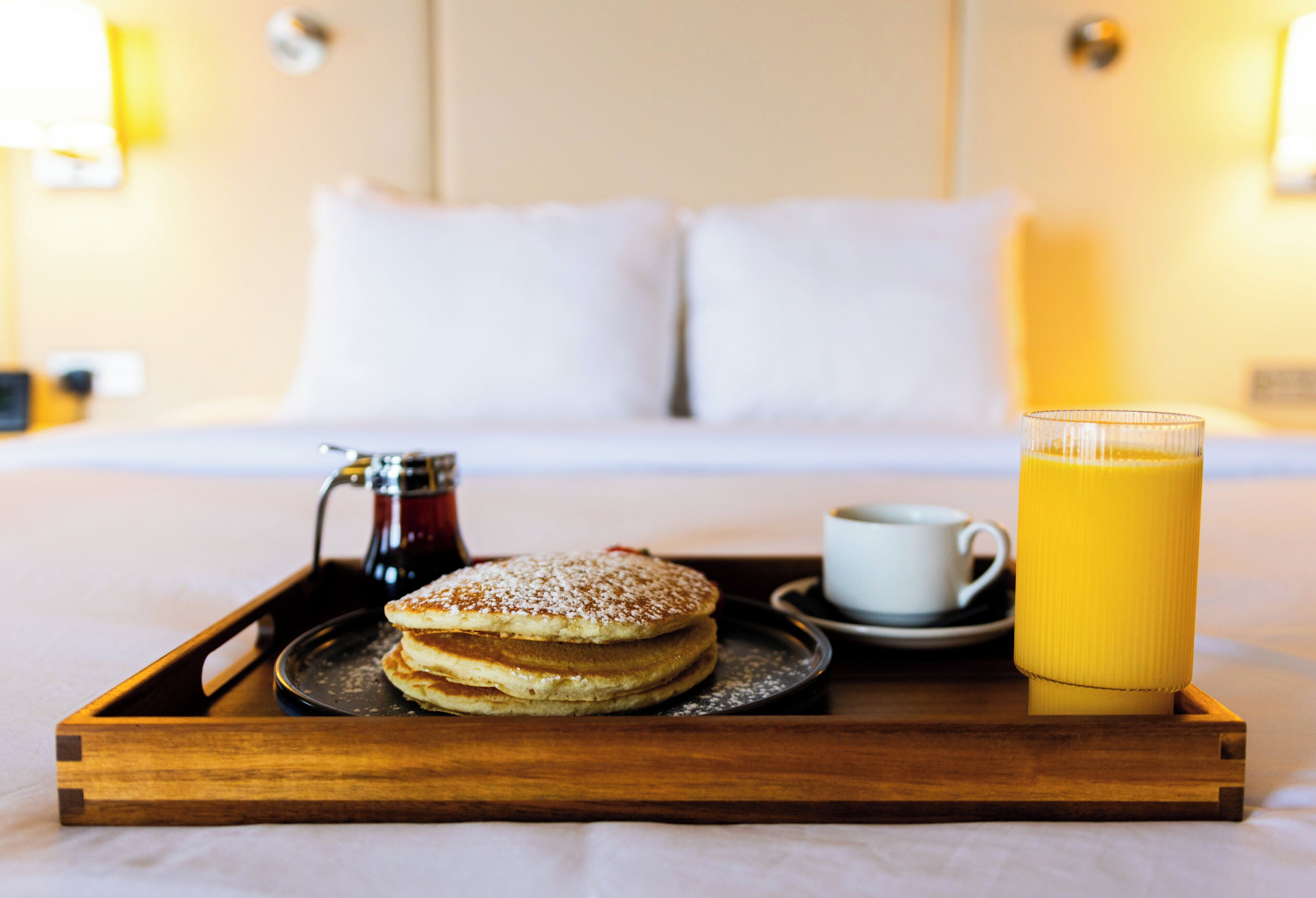 Un plateau de petit-déjeuner avec des crêpes, une tasse de café et un verre de jus d’orange, posé sur un lit.