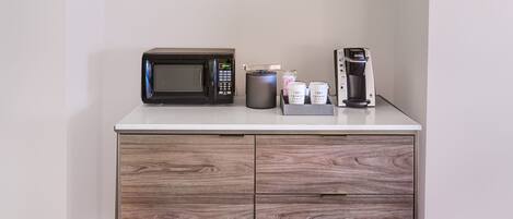Uma cozinha compacta com micro-ondas, cafeteira e cápsulas de café em uma bancada branca.
