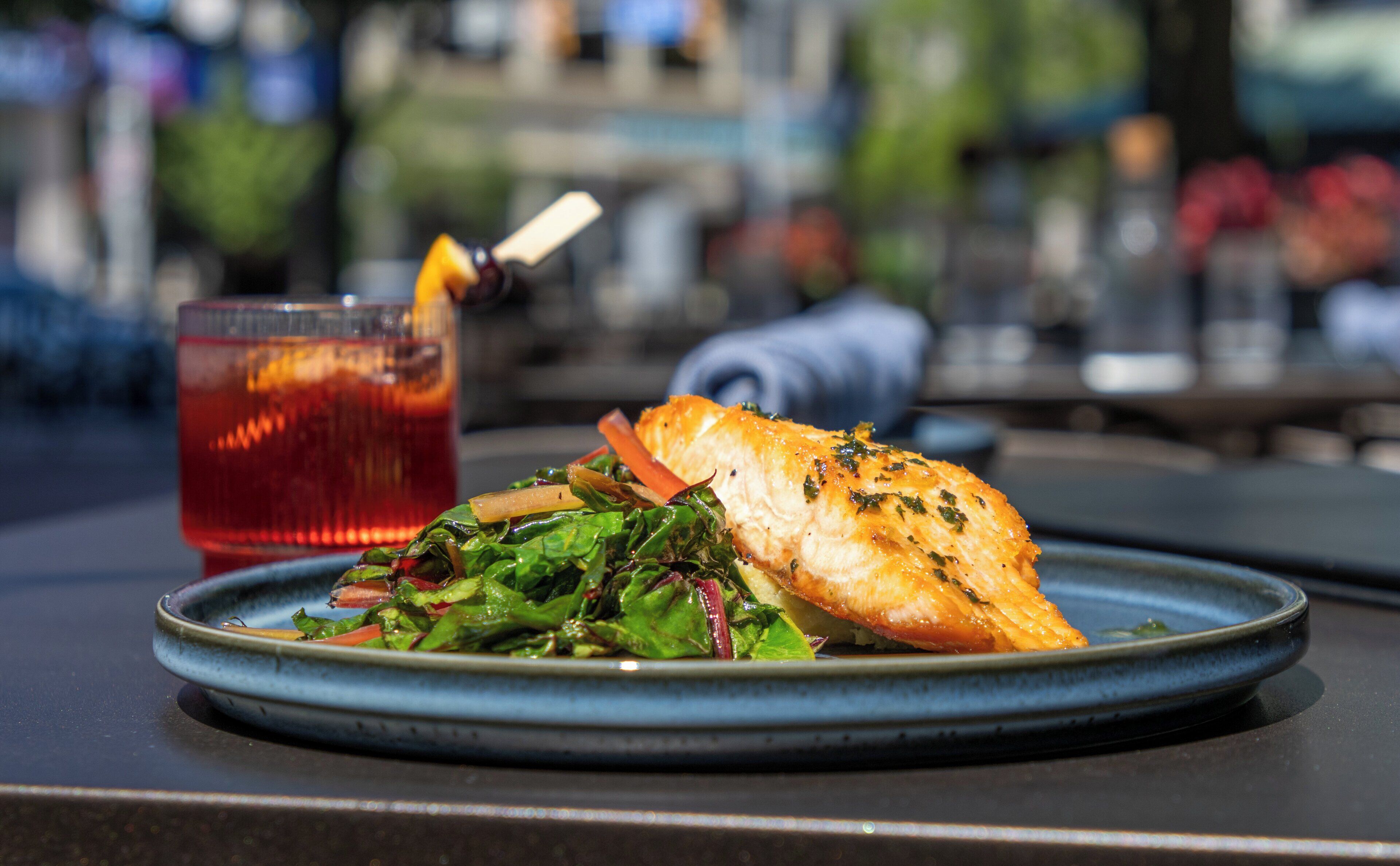 A plate with a piece of fish and greens, and a glass of red beverage with a lemon slice on a table.