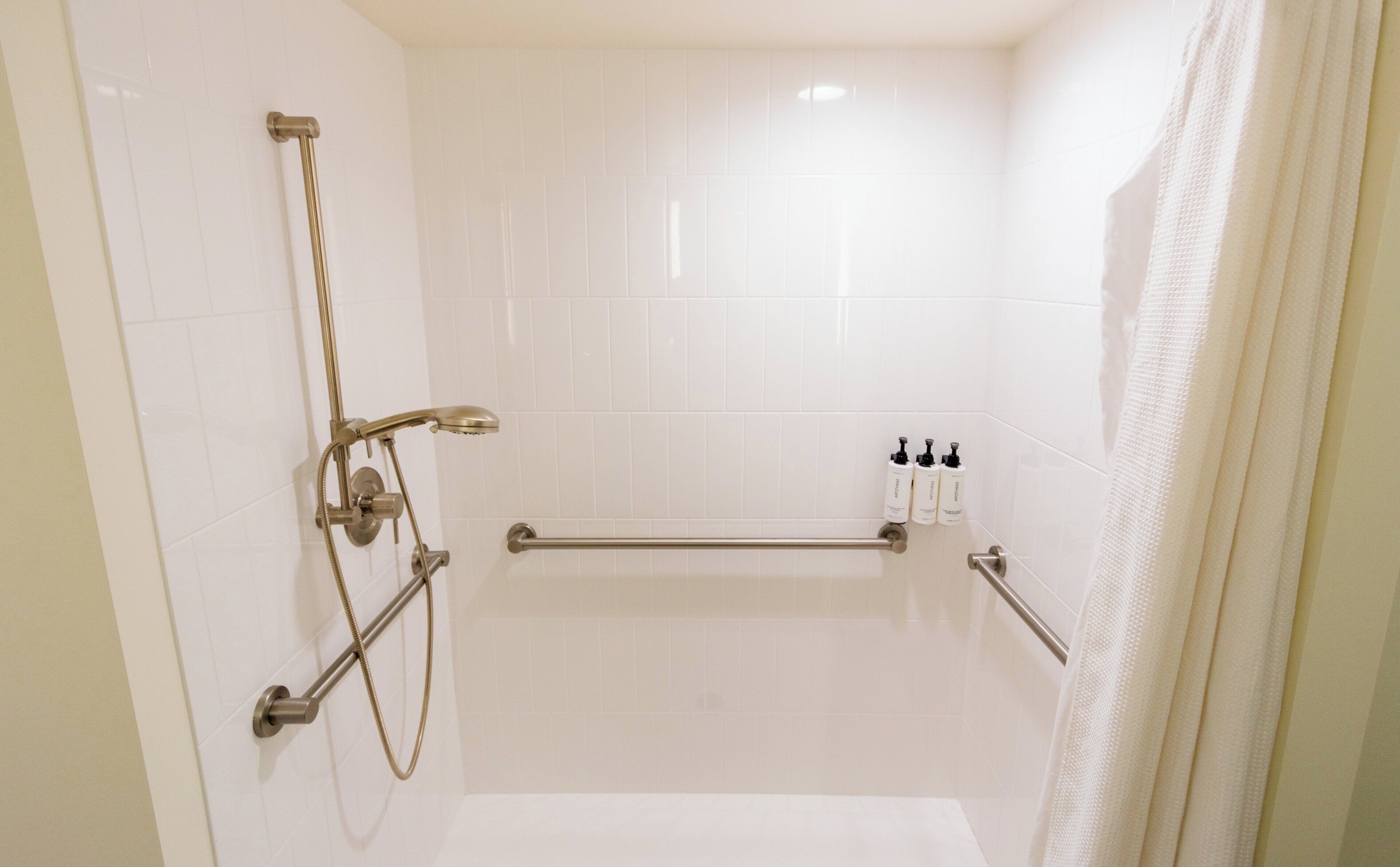 A modern bathroom with white tiles, a shower with a handheld showerhead, and a grab bar.