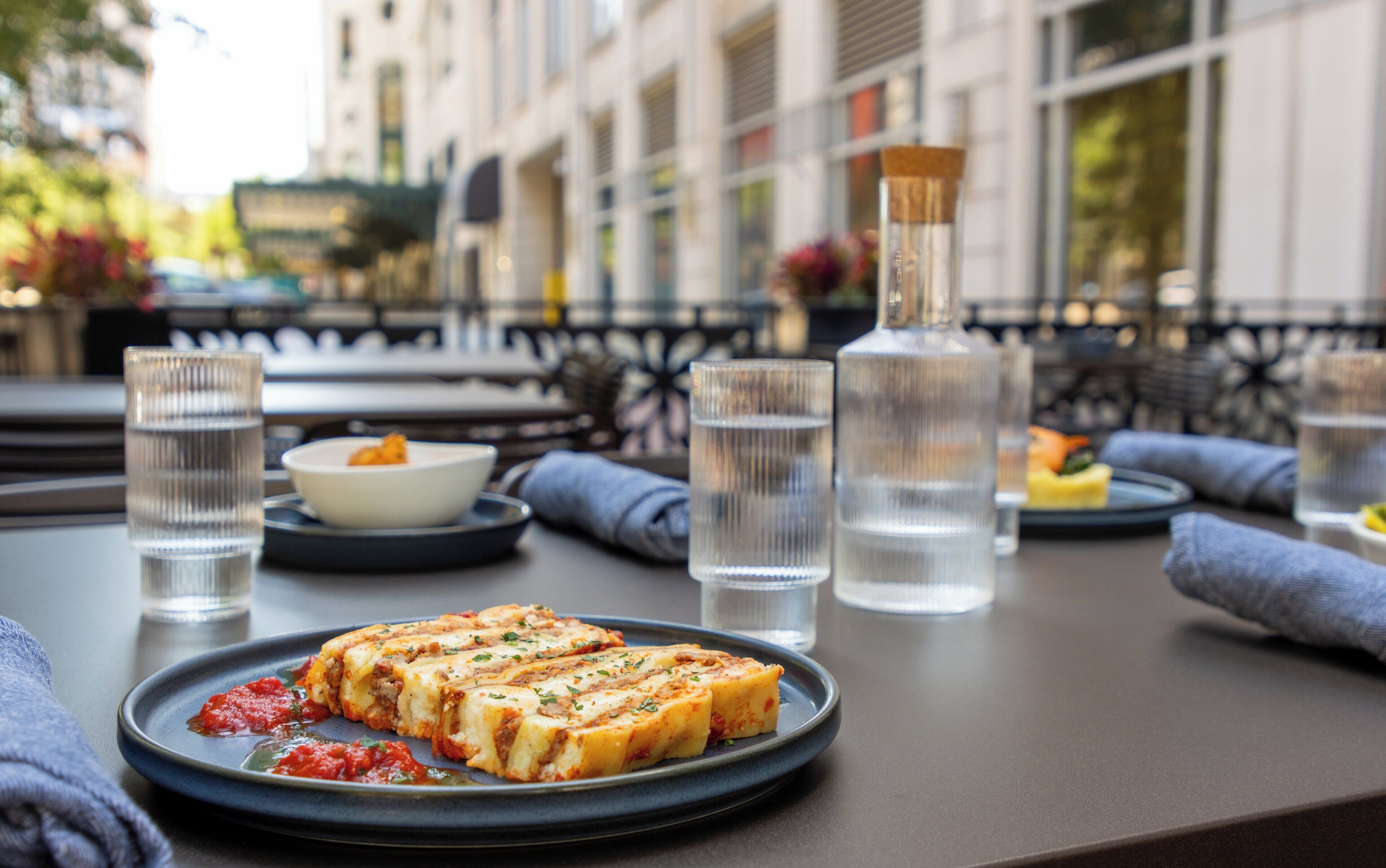 A table with a plate of food, glasses of water, and a bottle on a terrace with a view of buildings.