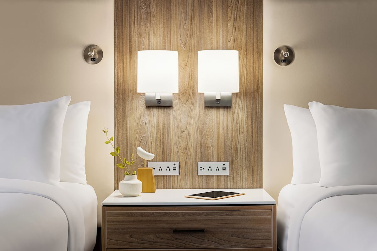 A hotel room with two beds, a bedside table with a lamp, a vase with a plant, and a tablet.